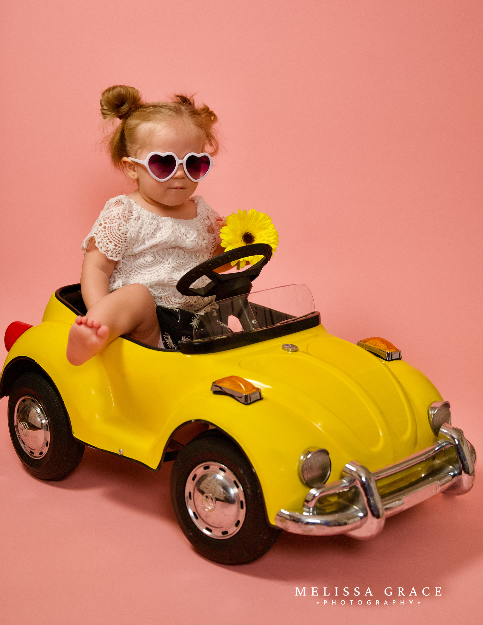 A young girl sitting in a yellow toy car with heart-shaped sunglasses, a white lace top, and holding a yellow flower, against a pink background.