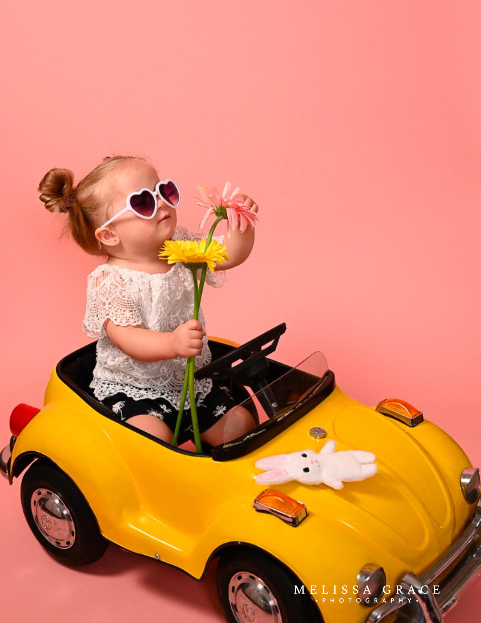A young girl with red hair styled in buns, wearing heart-shaped sunglasses and a lace white top, is sitting in a yellow toy car holding pink and yellow flowers. A white stuffed bunny toy is on the car's hood and the background is pink.