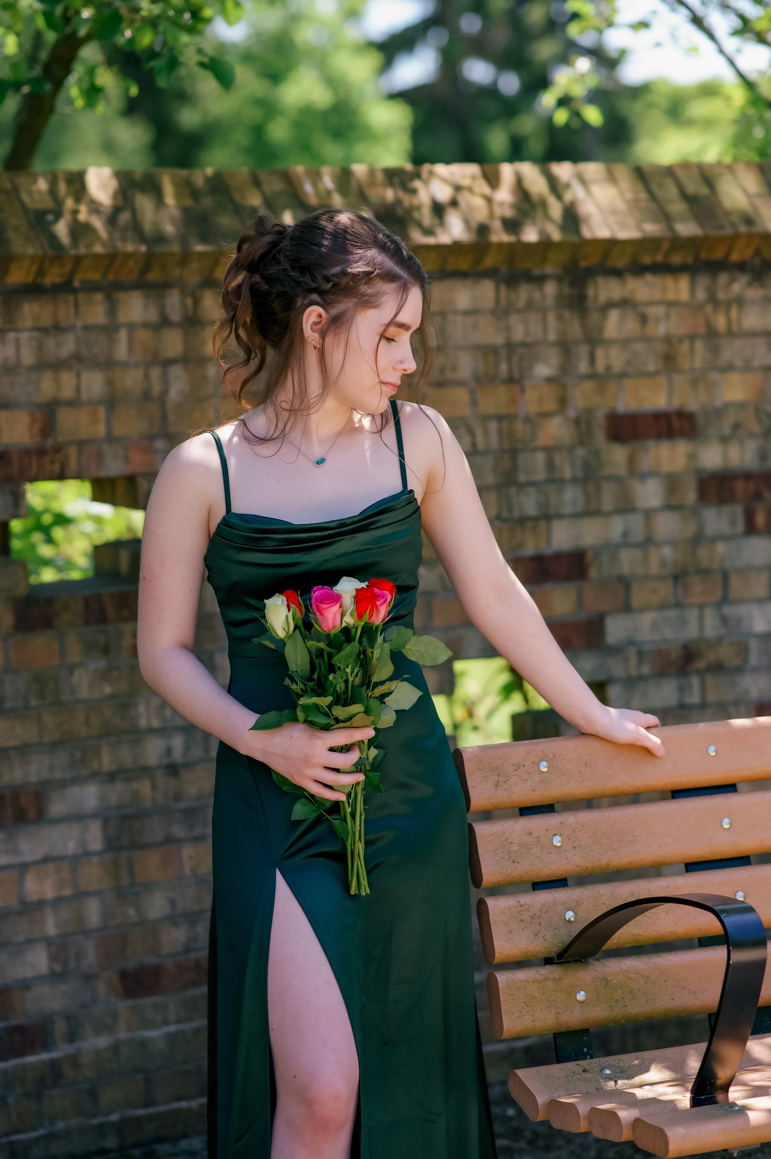 Young woman in a black dress with a high slit holding a bouquet of roses, standing outdoors near a bench and brick wall, with trees and greenery in the background.