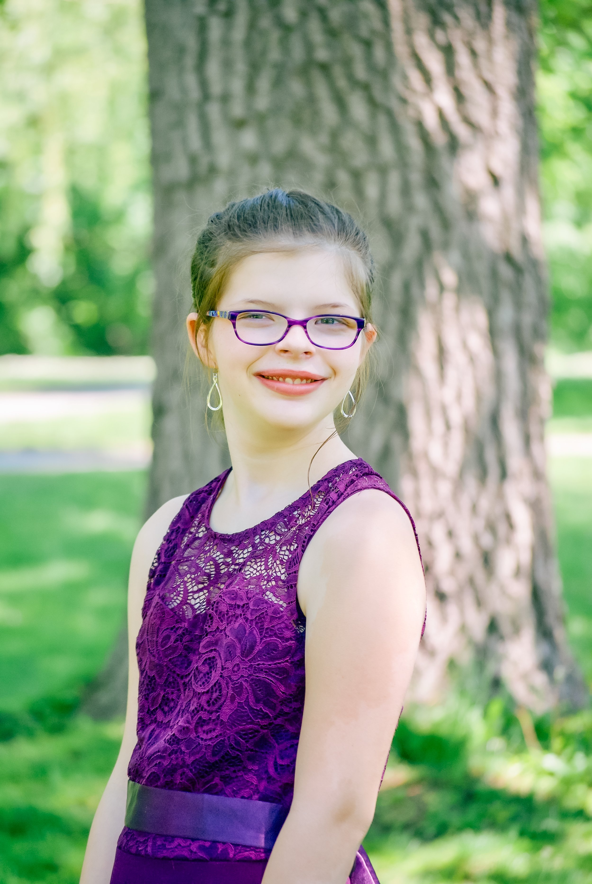 A young woman with glasses and earrings posing outdoors with a large tree in the background, wearing a purple lace dress.