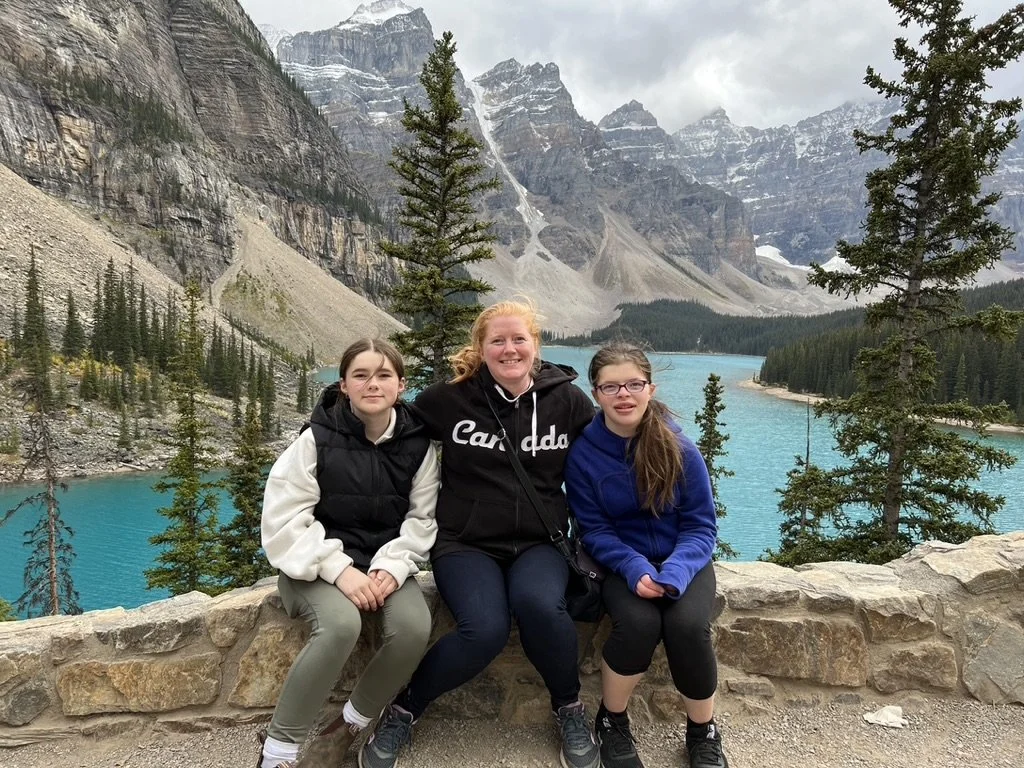 Three women sitting on a stone wall in front of a turquoise lake surrounded by mountains and evergreen trees.