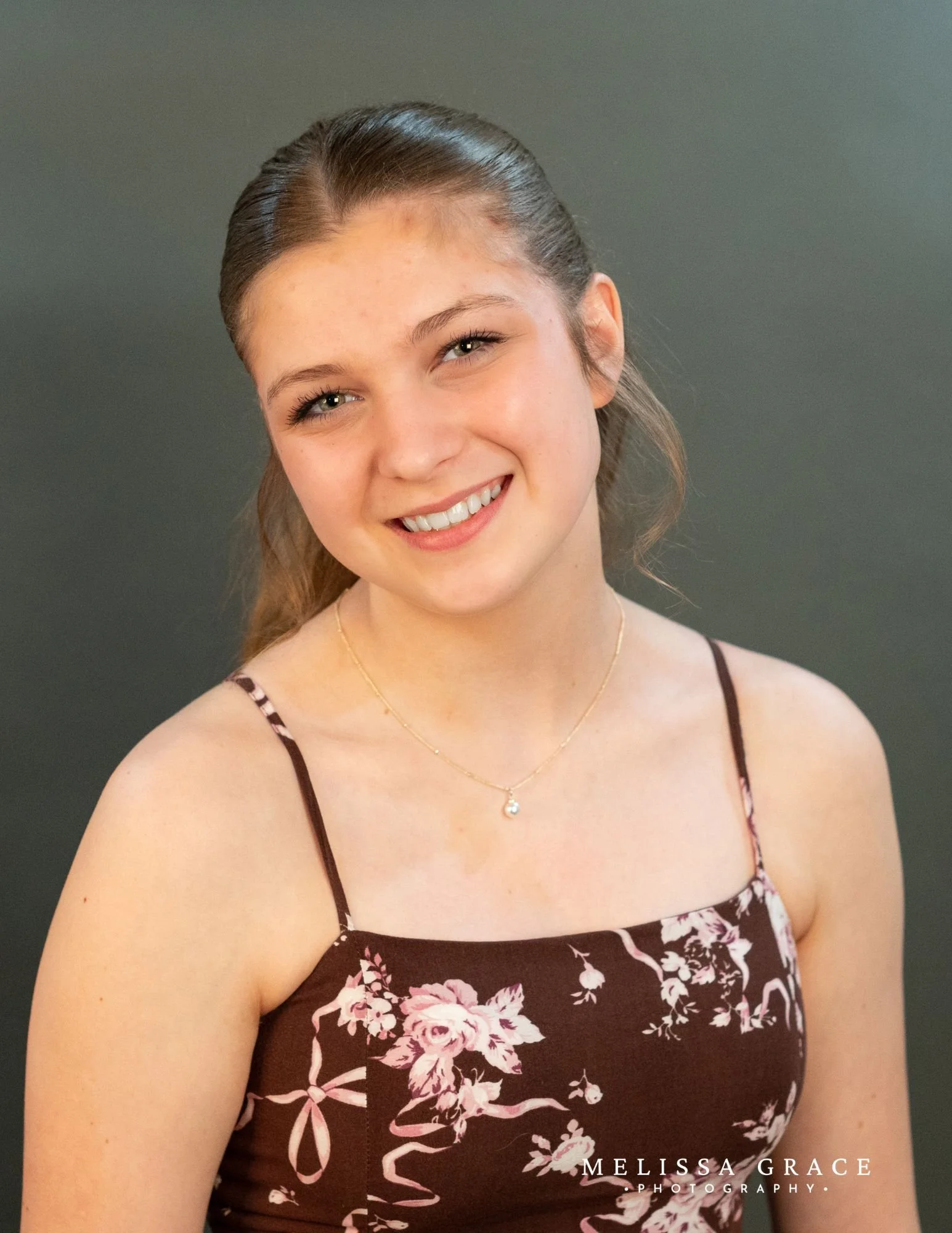 A young woman with light brown hair, styled with a side part, smiling with white teeth, wearing a dark brown floral dress with pink and white flowers, thin straps, and a gold necklace with a small pendant. The background is a plain, neutral color.