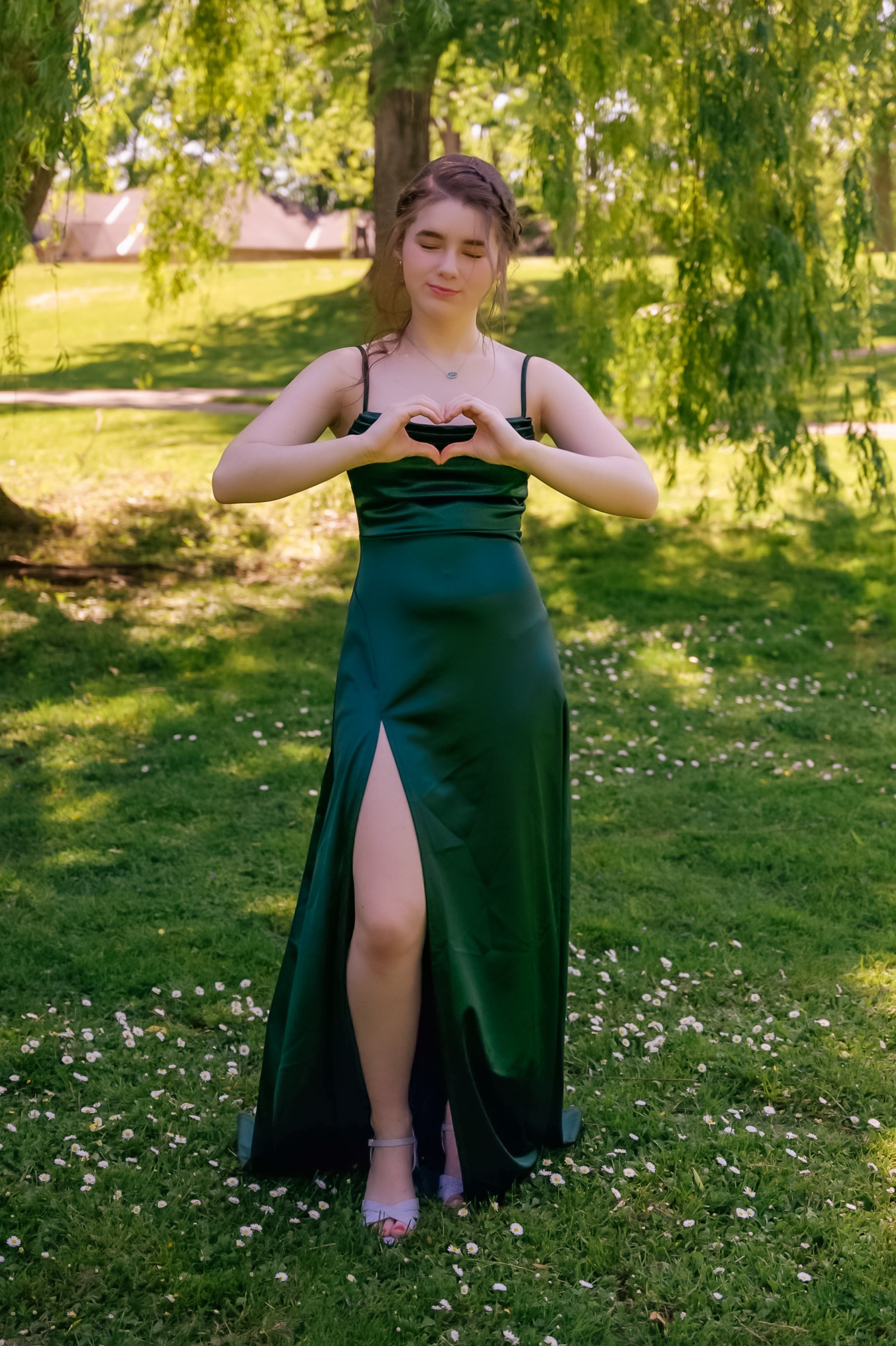 A young woman standing on a grassy field covered with small white flowers, wearing a black dress with a high slit, making a heart gesture with her hands, in a park with trees and a house in the background.