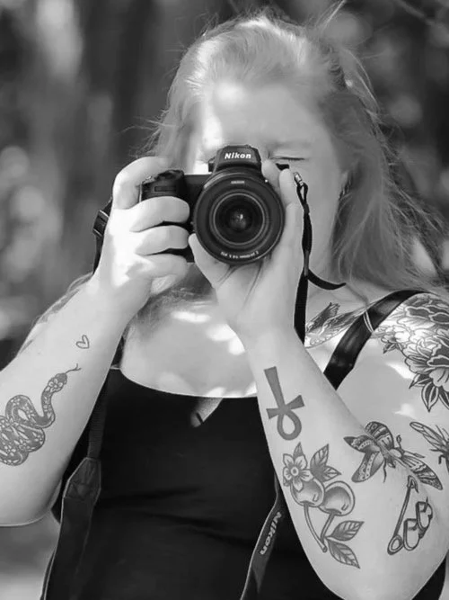 Person with tattoos holding a Nikon camera, taking a photo, outdoors in the sunlight.