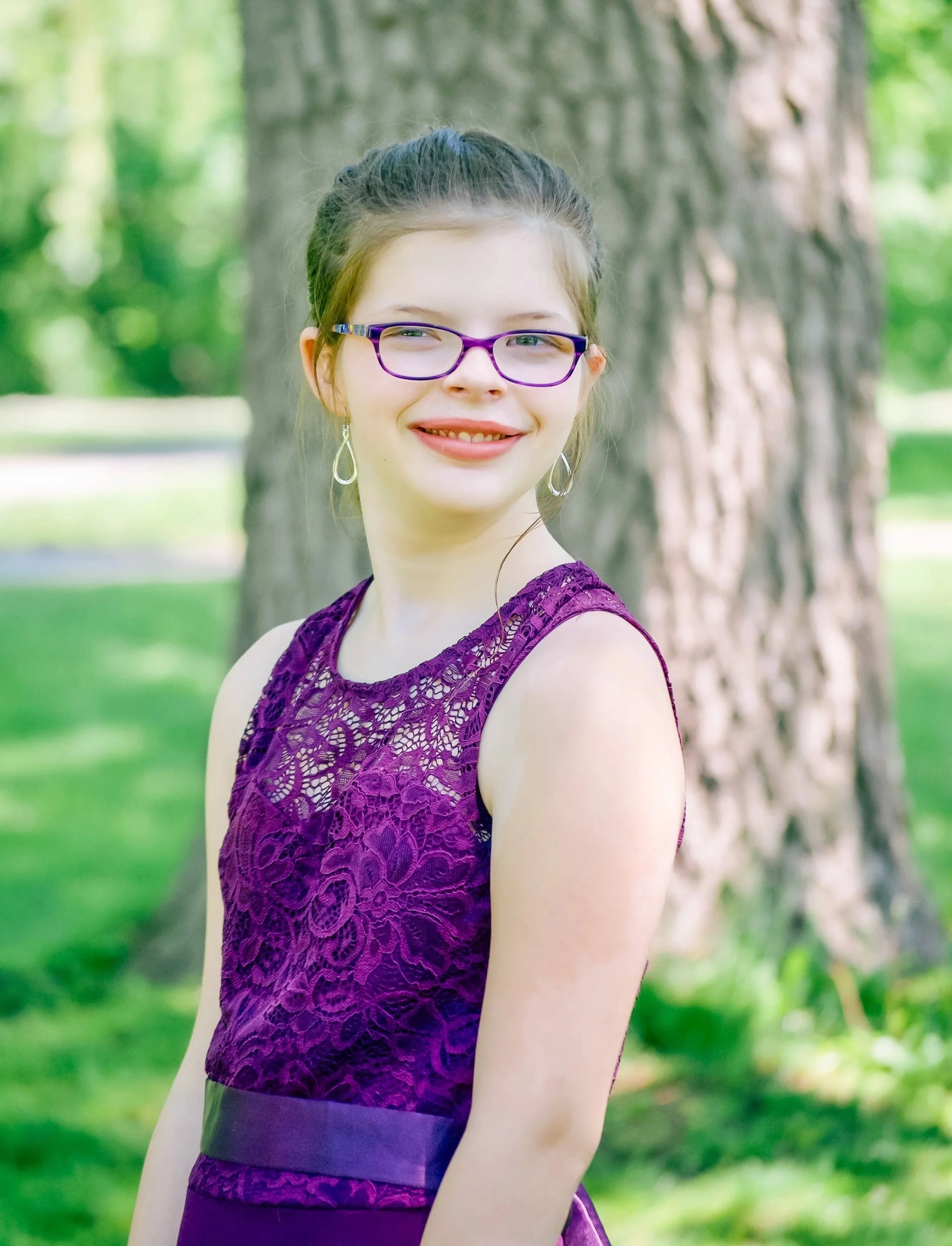 A young woman with glasses and earrings smiling outdoors in front of a large tree and green foliage.