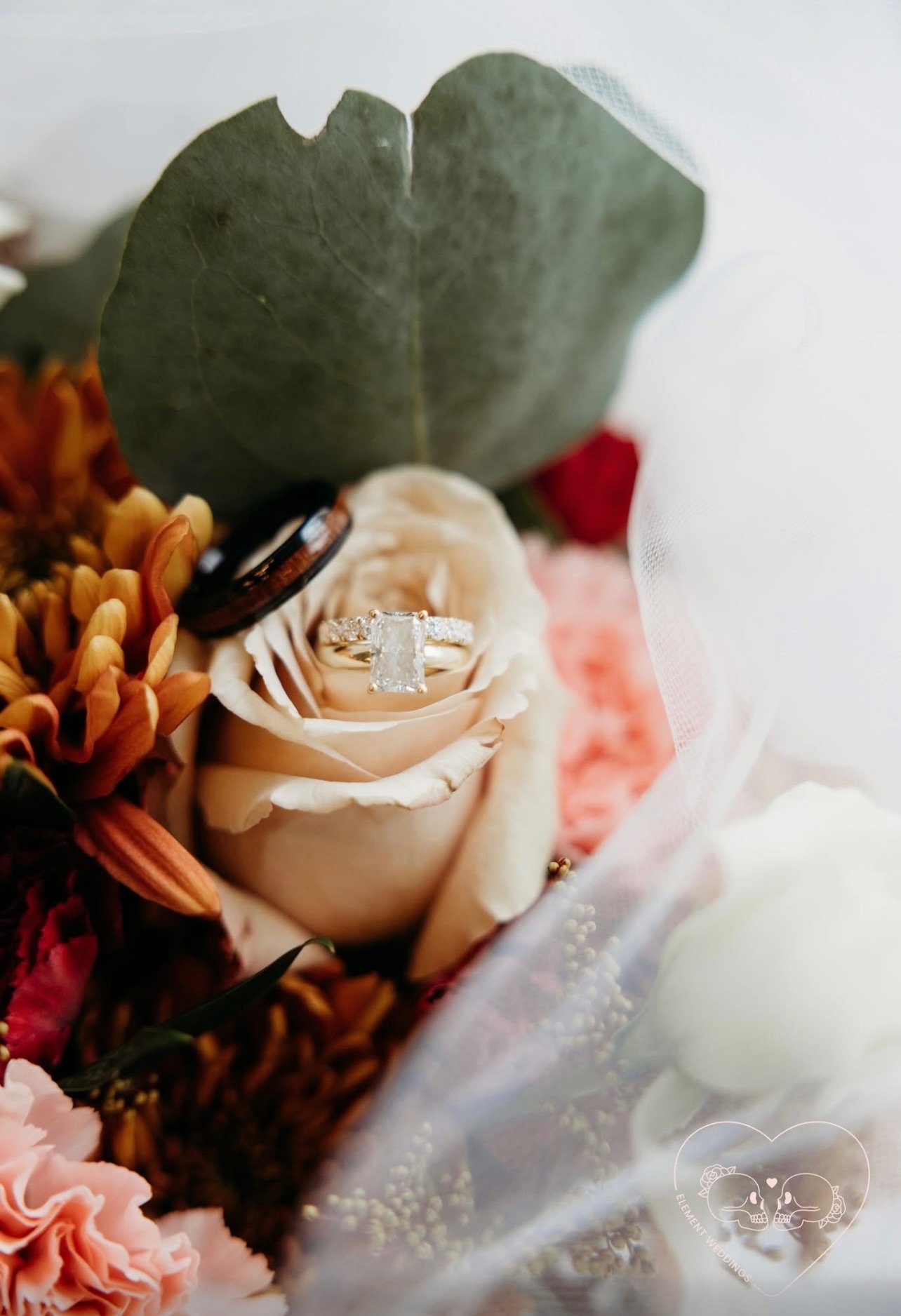 Close-up of an engagement ring with a large rectangular diamond, placed on a cream-colored rose, surrounded by orange and red flowers and green leaves, wrapped in sheer white fabric.