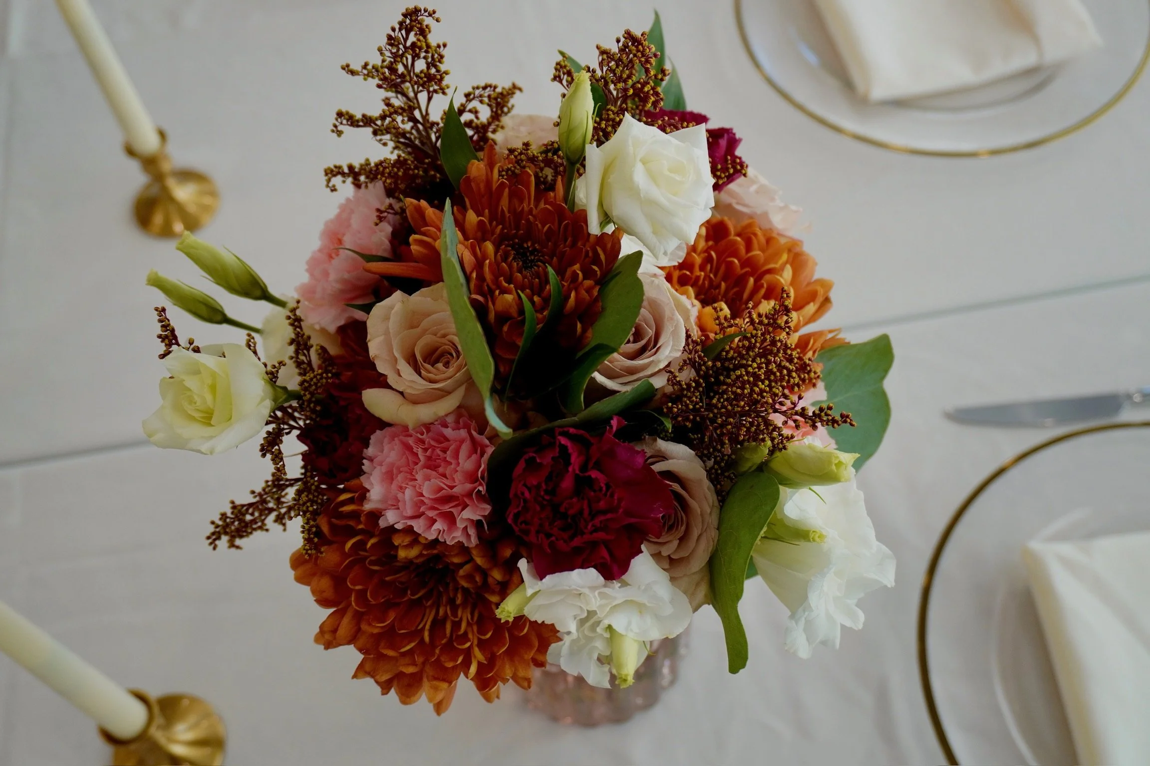 A floral arrangement in a pink vase with white, pink, orange, and red flowers, including roses, dahlias, and other blooms, on a table with a white tablecloth and gold candle holders.