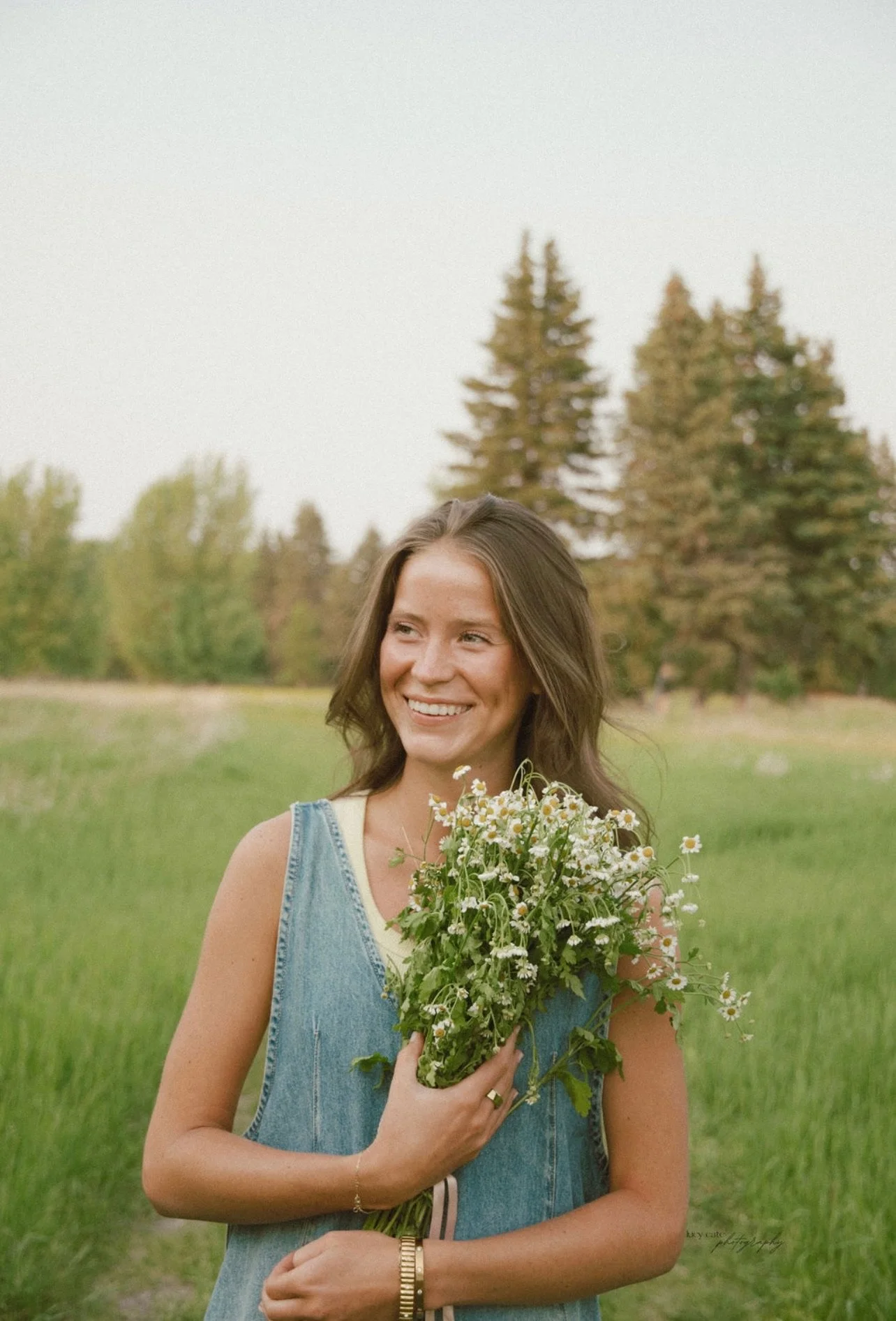 A woman with brown hair smiling, holding a large bouquet of daisies and wildflowers outdoors in a grassy field with trees in the background.