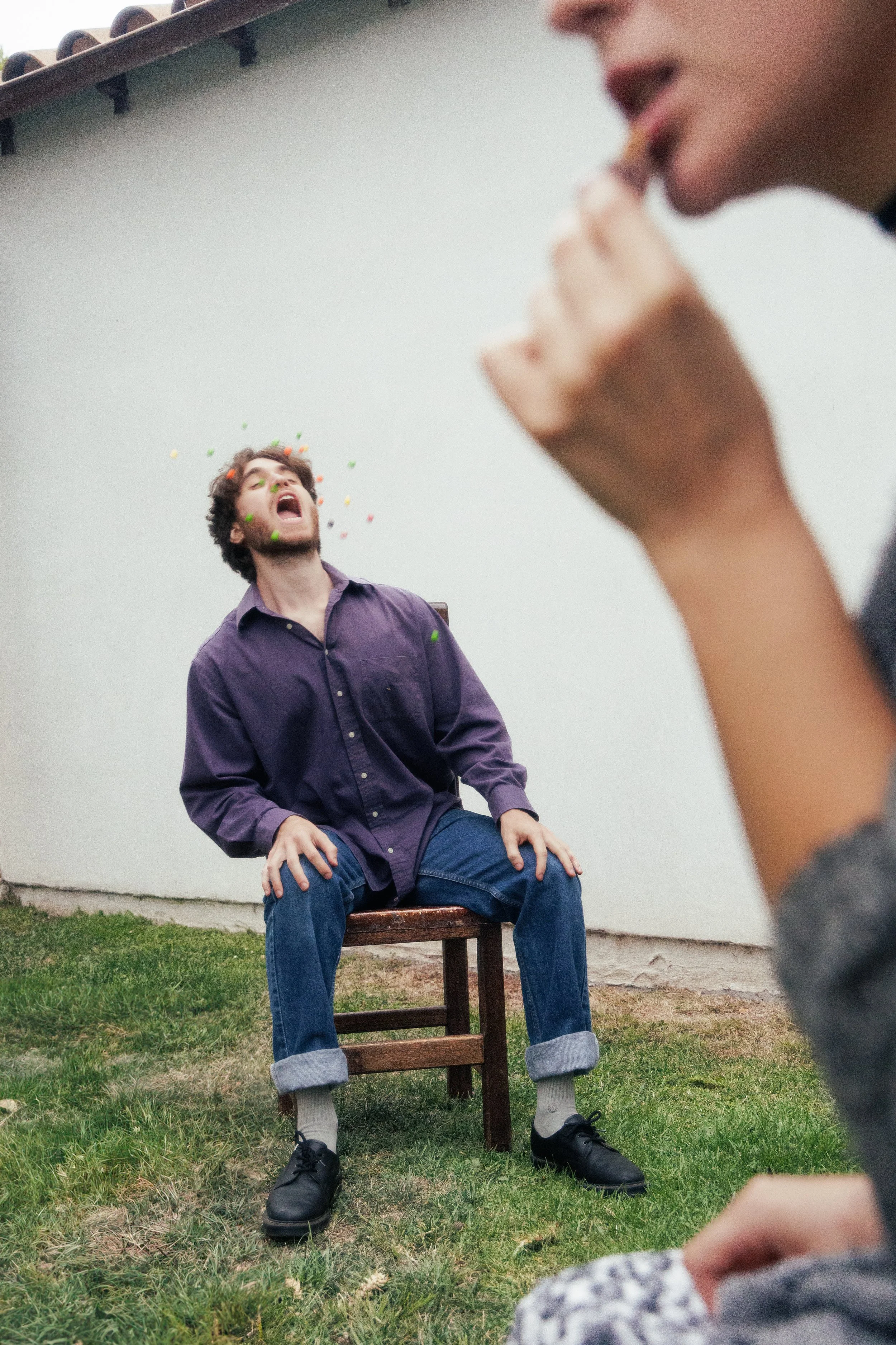 A man with curly hair and a beard is sitting on a wooden chair outdoors, leaning back with his head thrown back, shouting or laughing, while colorful confetti falls around him. Another person, partially visible, is in the foreground, raising a hand toward the man.