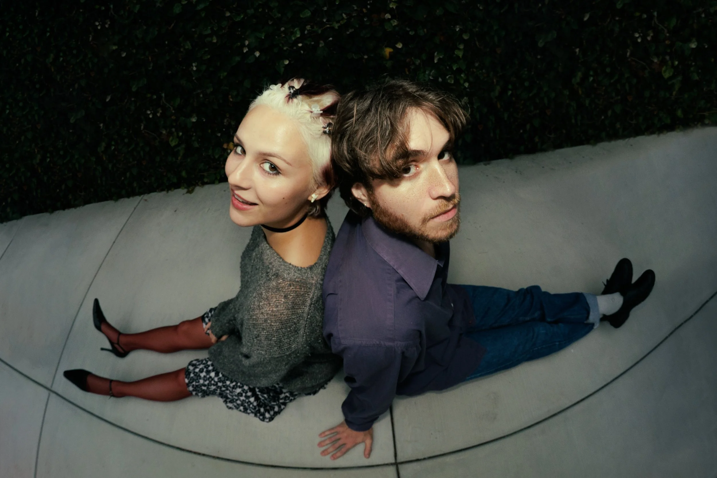 A woman and a man sitting back-to-back on a tiled floor outdoors, with a green hedge behind them. The woman has platinum blonde hair with hair clips, wears a gray sweater, black high heels, leopard-print pants, and a black choker. The man has brown hair with a beard, wears a purple shirt, blue jeans, and black shoes. They look up at the camera over their shoulders.