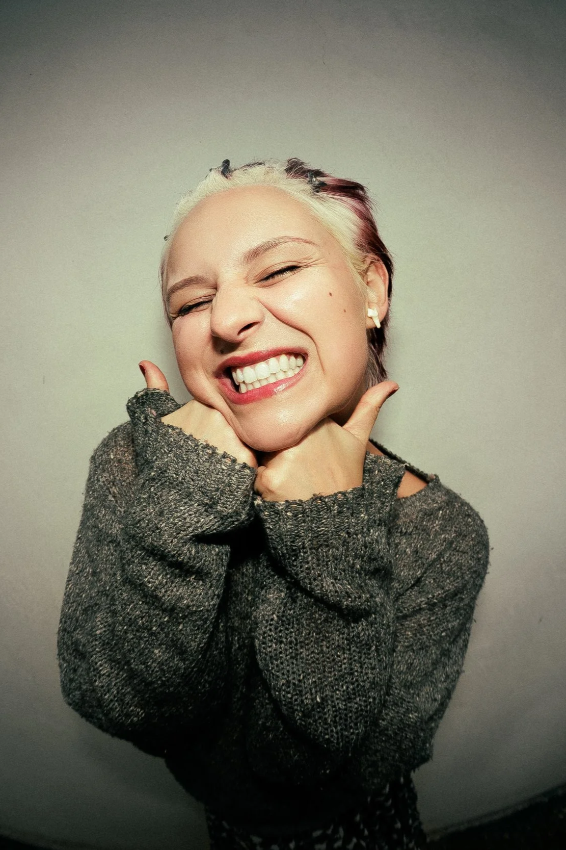 A woman with blonde and red hair smiling widely with eyes closed, pressing her cheeks with her hands, wearing a gray sweater.