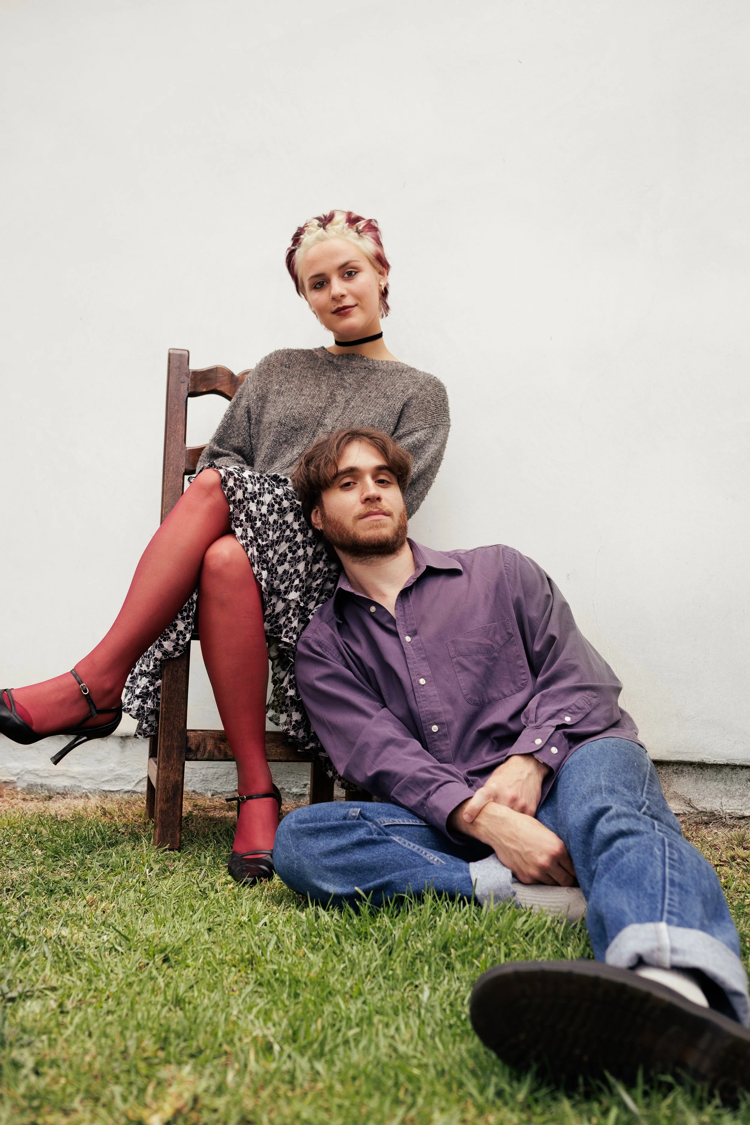 A young woman with short, multicolored hair and a young man with brown hair are sitting together outdoors against a plain white wall. The woman is sitting on the man’s lap, wearing a gray sweater, a floral skirt, red stockings, and black heels, and looking at the camera. The man is lying on the grass, wearing a purple shirt and blue jeans, with his head resting against the woman's leg, looking at the camera.