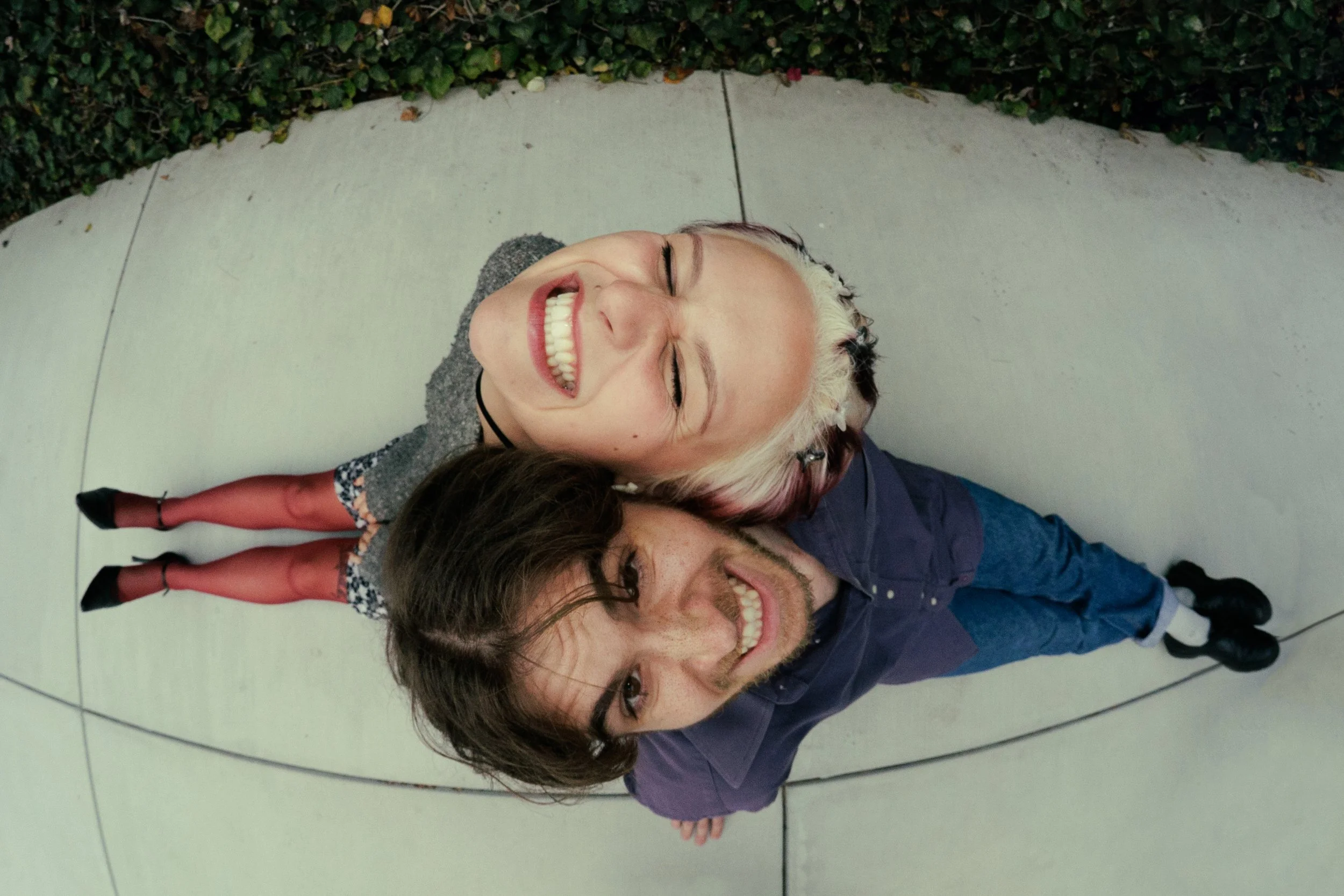 Two people lying on the floor side by side, smiling and appearing happy. They have dark and light hair, respectively. Behind them is a person with black hair facedown, wearing black high heels and patterned socks. The background includes a green hedge and a tiled floor.