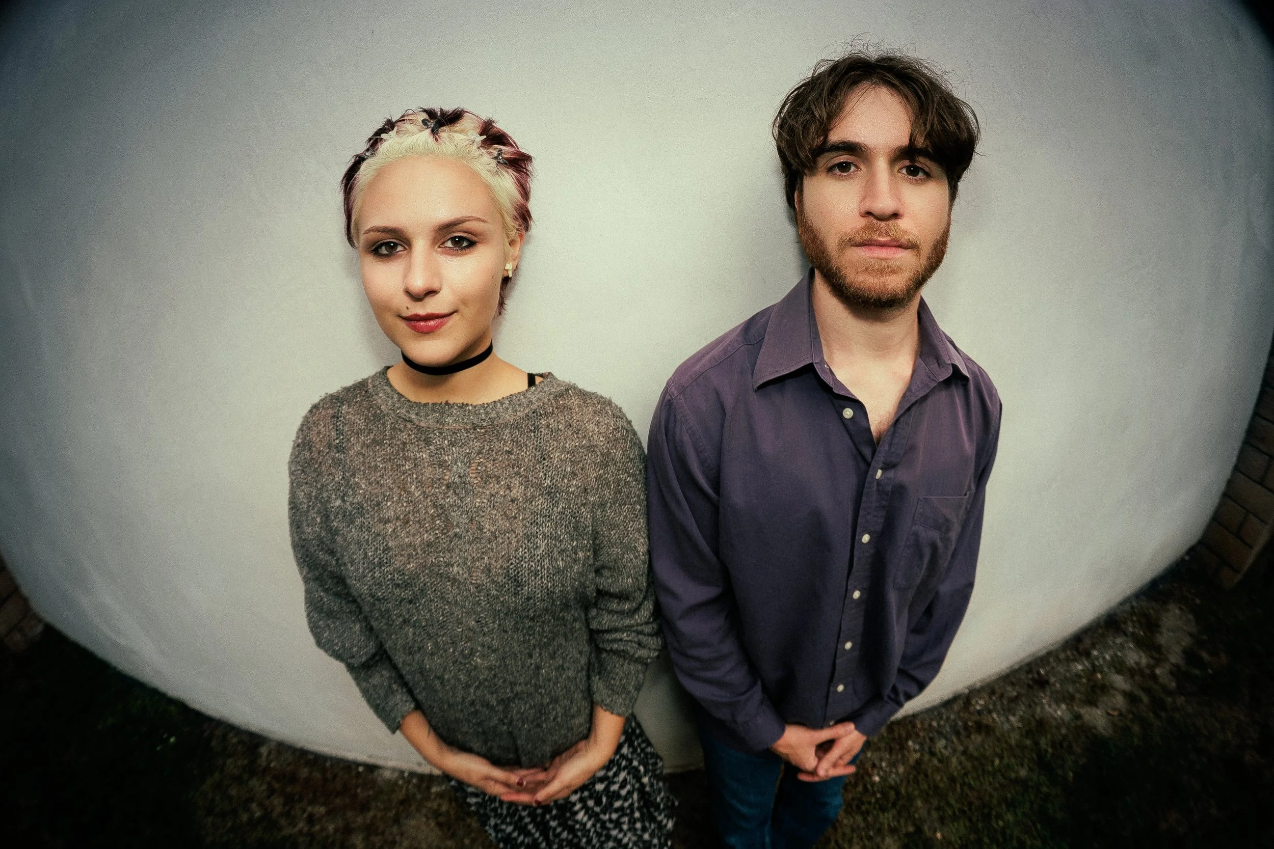A young woman with short, multicolored hair and a young man with brown hair and a beard stand side by side against a plain wall, looking at the camera.