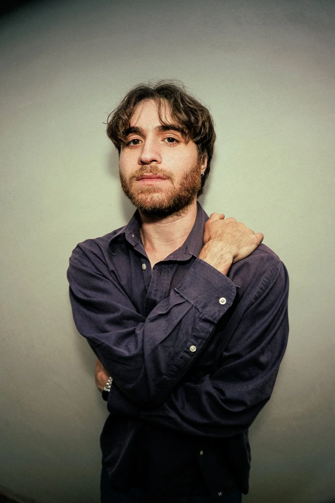 A man with brown hair and a beard wearing a dark purple button-up shirt, standing against a plain wall, with one hand touching his shoulder and looking at the camera.