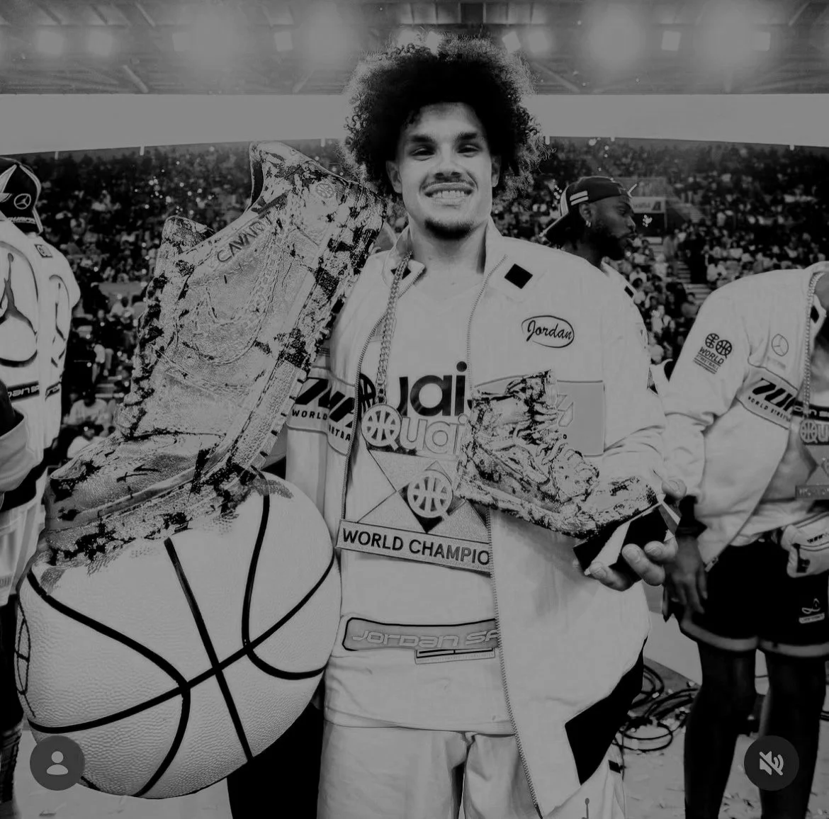 A smiling man in a sports uniform holding a basketball and a trophy in a stadium filled with spectators.