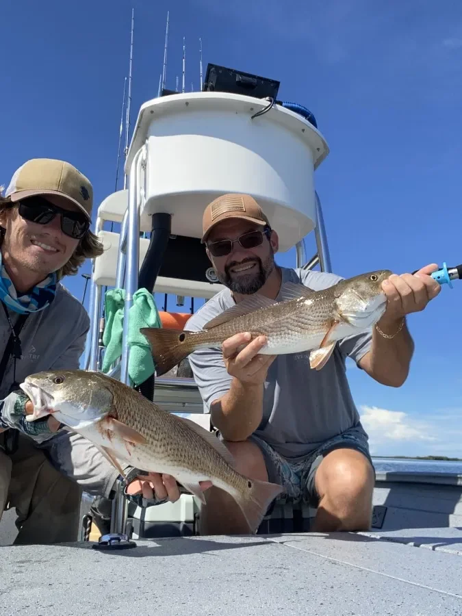 Two men on a boat holding fish they caught, with the boat's tower and antennas visible against a clear blue sky.