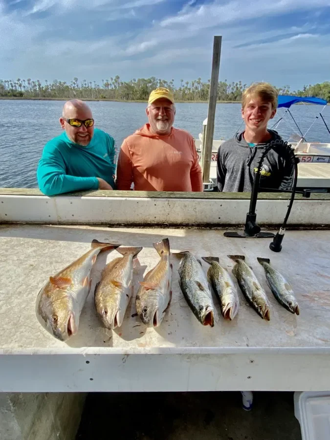 Three men standing behind a table with six fish caught in the water, on a boat docked by a lake with clear skies and trees in the background.