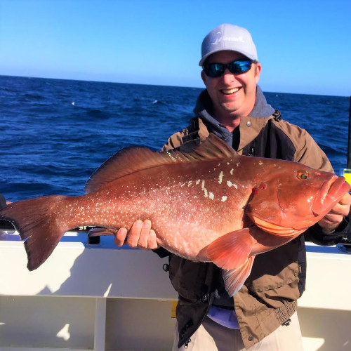 A smiling man on a boat holding a large fish he caught, with the ocean in the background during daytime.