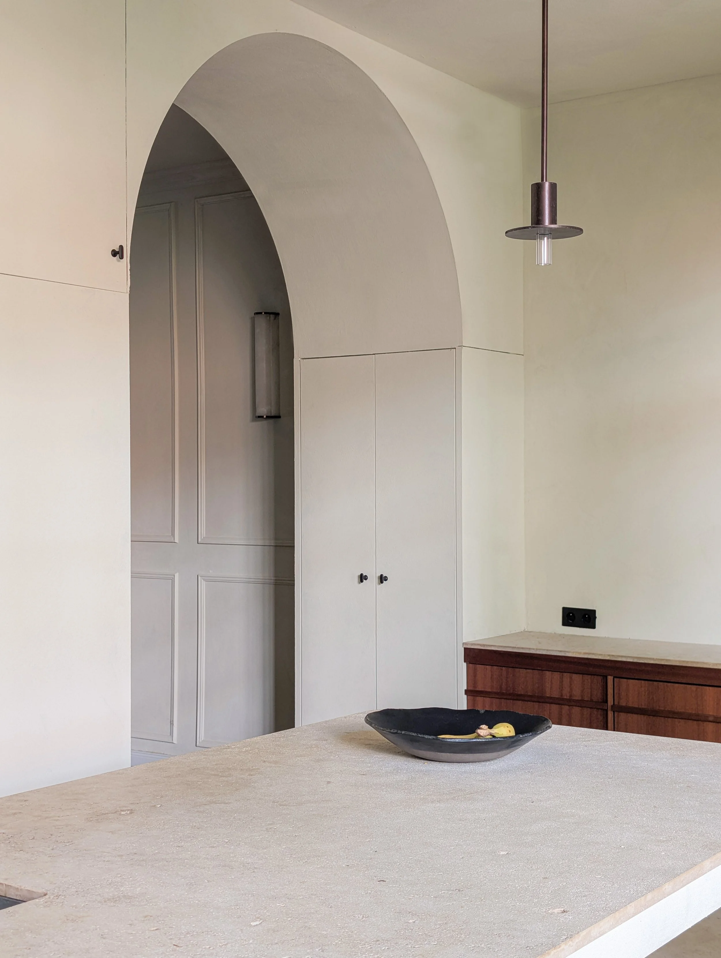 Een modern keukenblok met een beige werkblad, een zwarte kom met wat fruit, een houten lade onder het werkblad en een witte muur met een boog en kasten. Er is ook een lamp aan het plafond en een wandcontactdoos op de muur.
