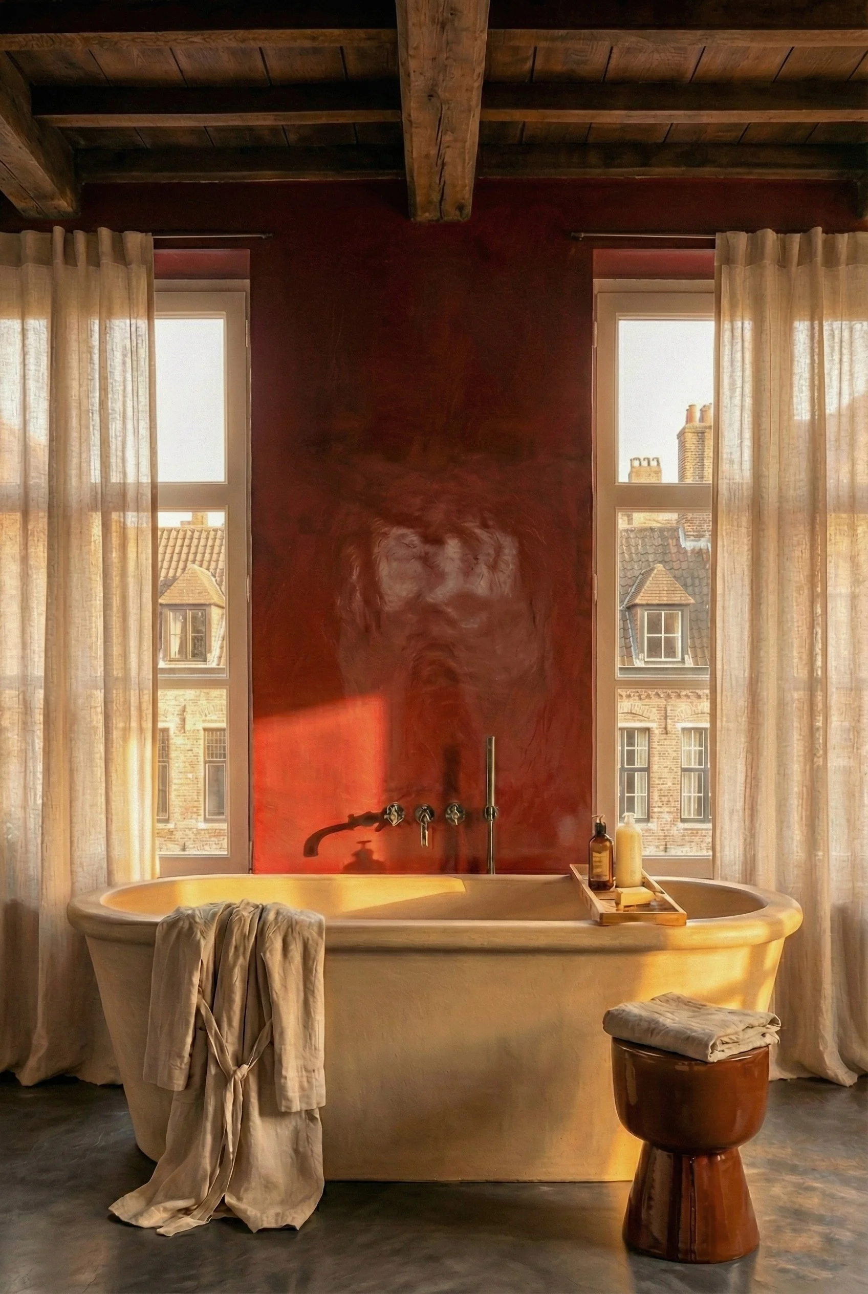 Een badkamer met een vrijstaand bad, gelegen voor grote ramen met beige gordijnen. Om het bad heen liggen handdoeken en badproducten, en er hangt een roodbruine muur met een houten plafond.