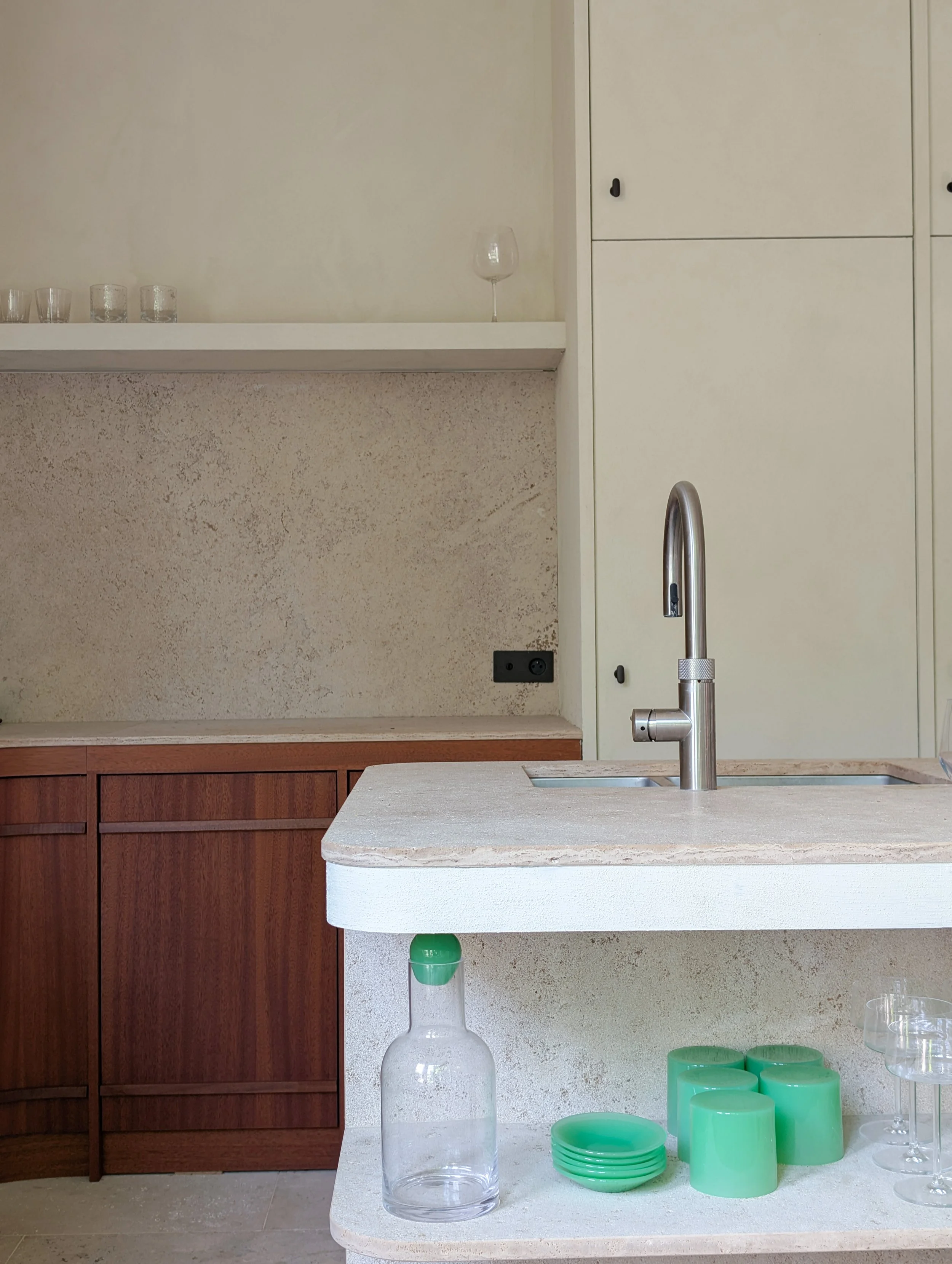 een keuken met een witte gootsteen, houten kasten en een beige muur. Op het aanrecht staan groene bekers, een grote fles met een open mond, en glazen. Op de muur hangen witte kasten en een witte plank met drie glazen, een vaas en een wijnglas.