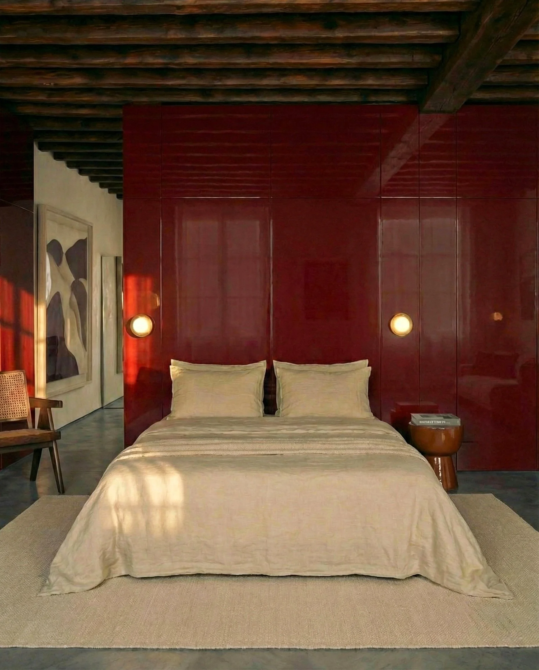 Een slaapkamer met een groot bed dat is bedekt met een beige dekbedovertrek en twee beige kussens. Aan weerszijden van het bed hangen twee ronde muurlichten, en rechts staat een ronde bijzettafel met een stapel boeken en een leeslamp. Aan de linkerkant hangt een kunstwerk met abstracte vormen.
