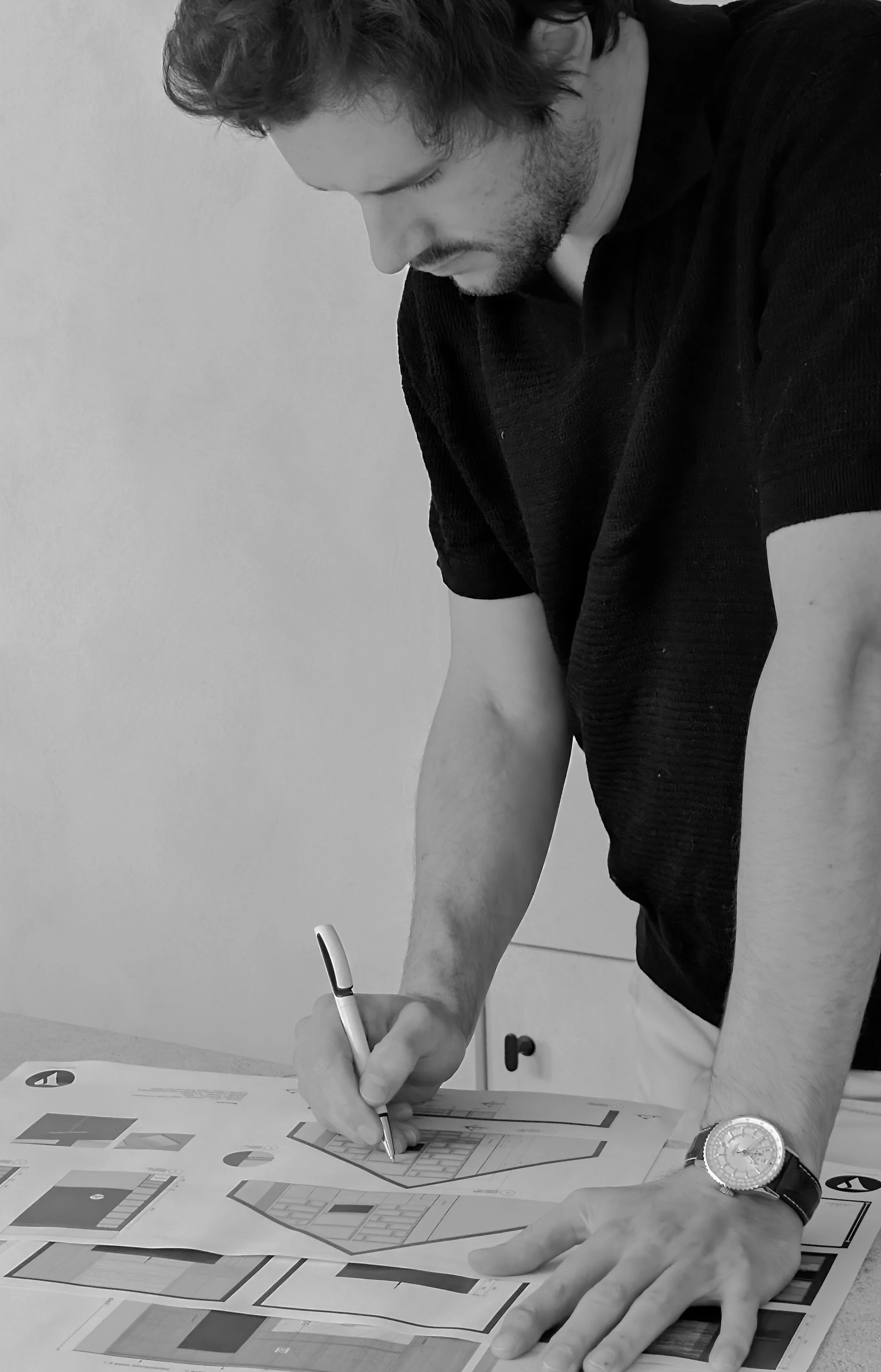 Man tekent op een plattegrond met een pen, terwijl hij op een tafel ligt, gekleed in een zwart T-shirt en een horloge op zijn linkerpols.