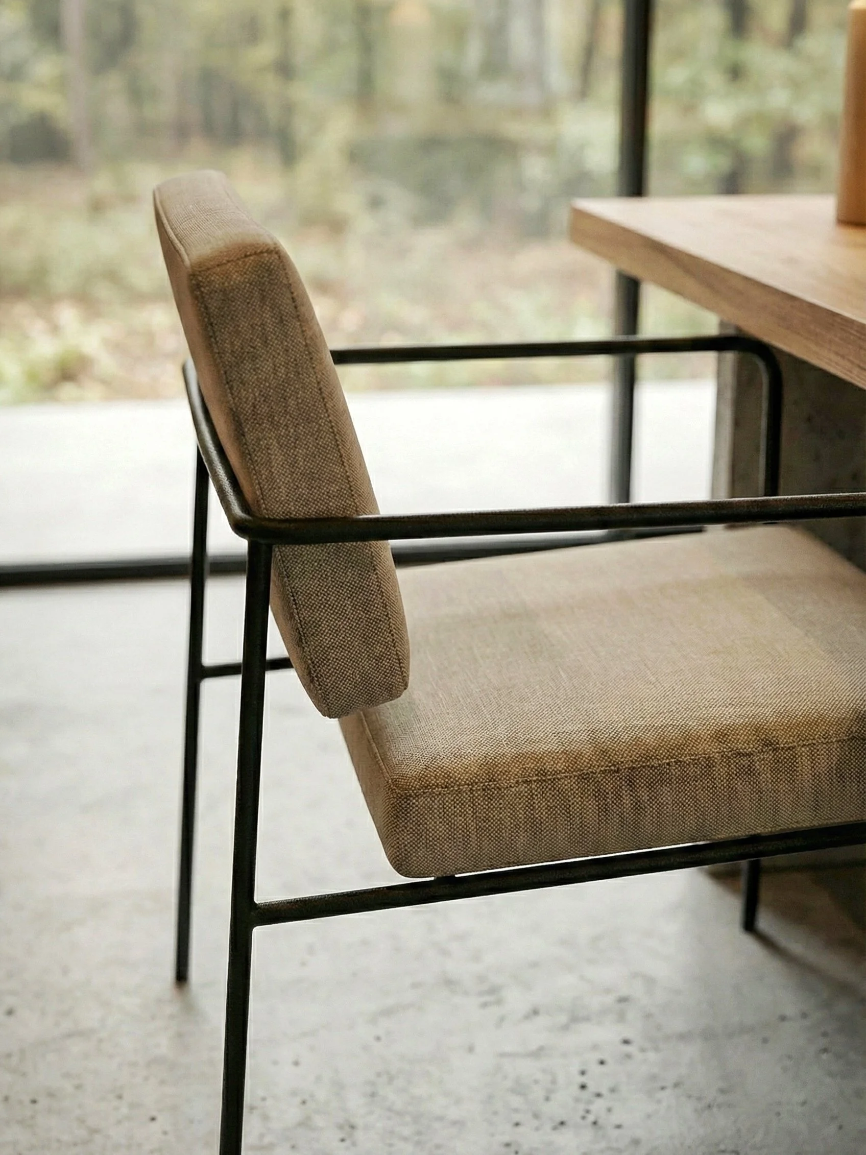 Een beige bureaustoel met een metalen frame voor een raam met uitzicht op een bos of park.