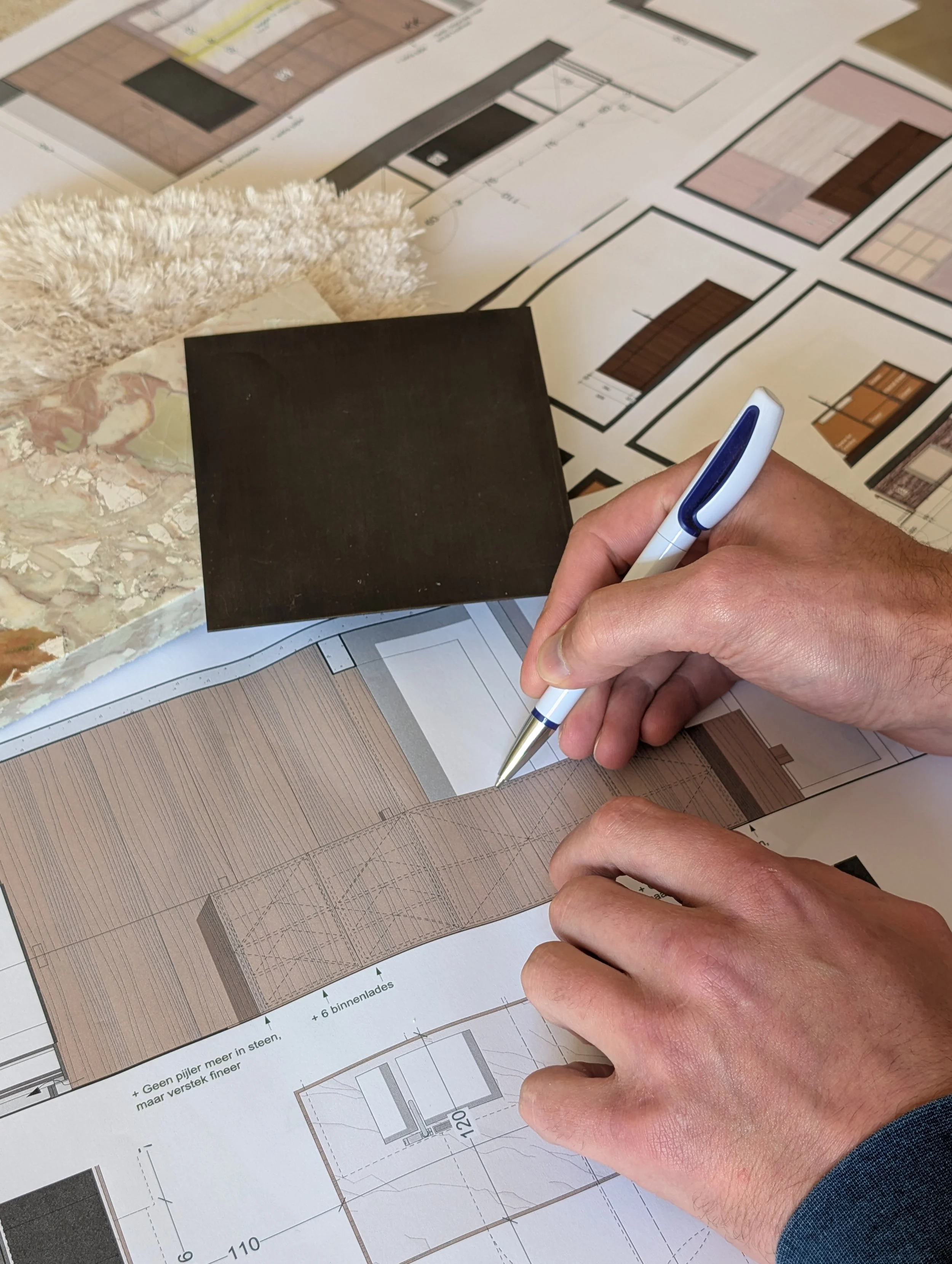 De afbeelding toont een persoon die een pen vasthoudt en werkt aan bouwplannen en plattegronden voor een bouwproject. Op de tafel liggen verschillende gedetailleerde documenten en kleurige constructie-ontwerpen.