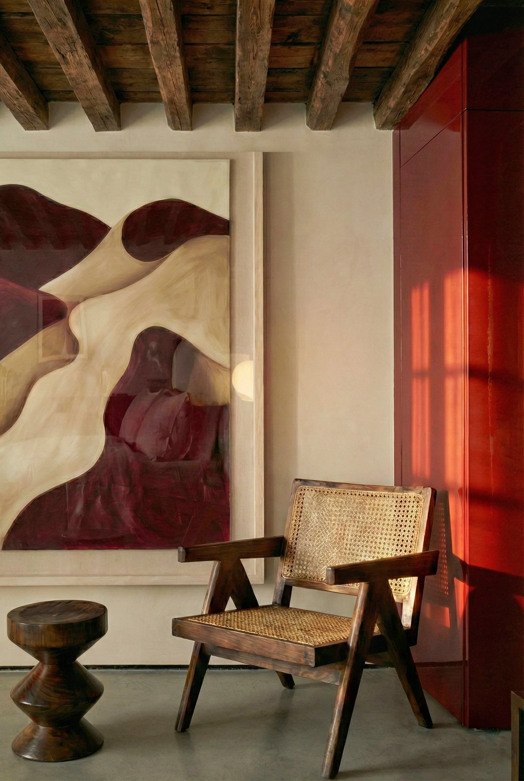 Een interieur met een houten plafond met zichtbare balken, een abstract schilderij met rode en beige tinten, een donkerrode kast, een houten stoel met een gevlochten zitting, en een kleine houten stool.