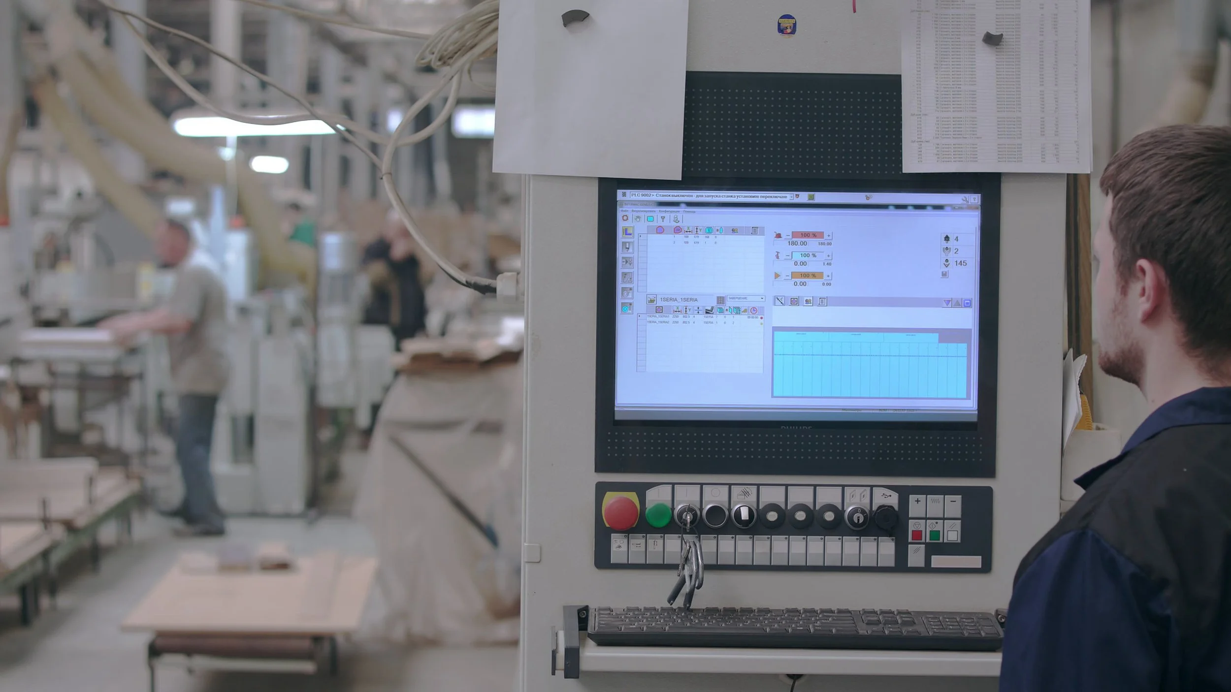 Een persoon in een fabriekshal kijkt naar een computerscherm dat een grafiek en gegevens toont, omringd door industriële machines en werknemers.