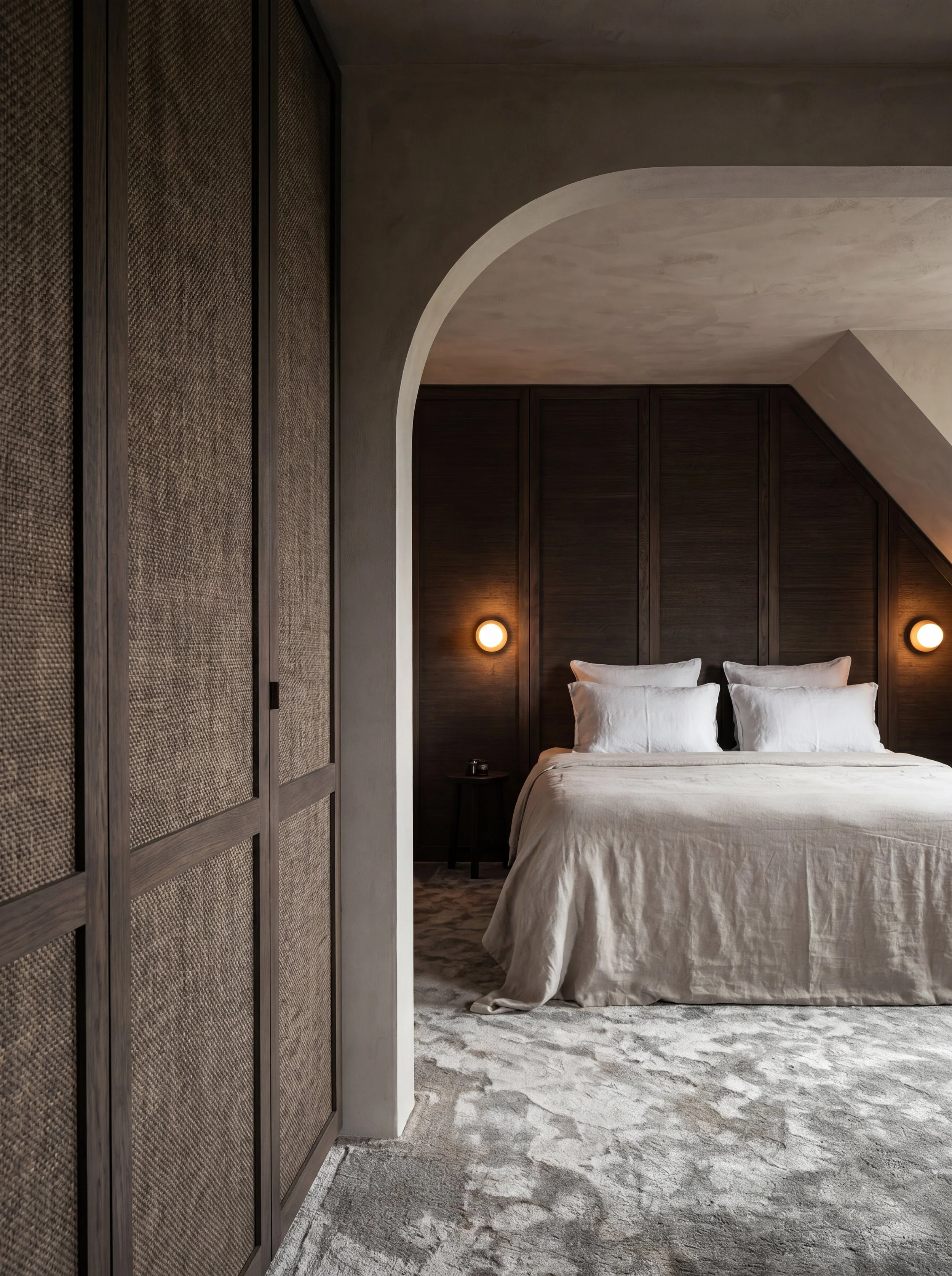 Een slaapkamer met een zacht beige beddengoed, donkere houten wandpanelen achter het bed, wandlampen aan weerszijden en een grote, gebogen witte muur. Aan de linkerzijde een kast met matarmaturen, het tapijt is licht met een patroon.