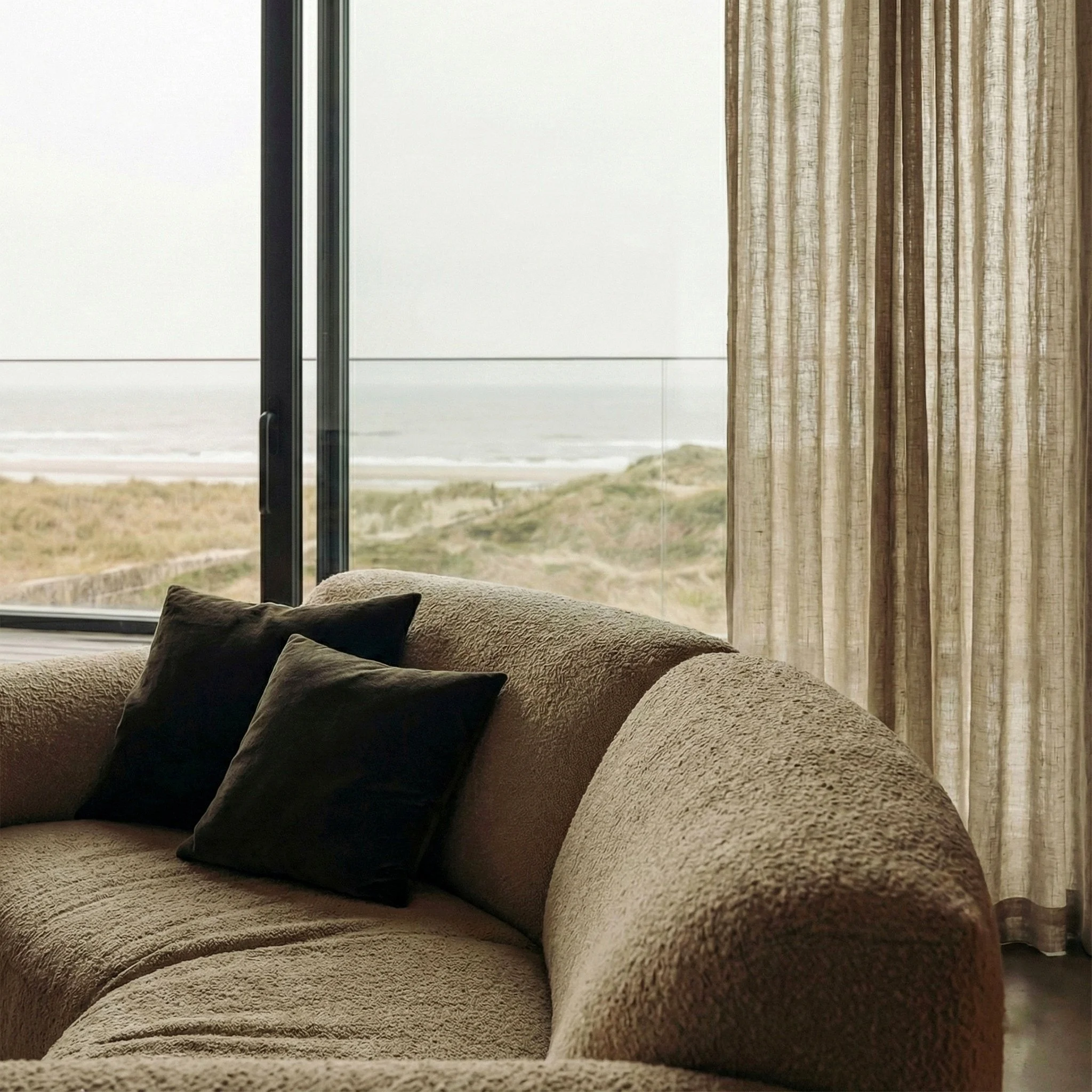 Binnenzicht van een woonkamer met een grote, beige, geruite bank en meerdere kussens in donkerbruine en beige kleuren. Achtergrond zichtbaar door raam, met een kustlijn en de oceaan in de verte.