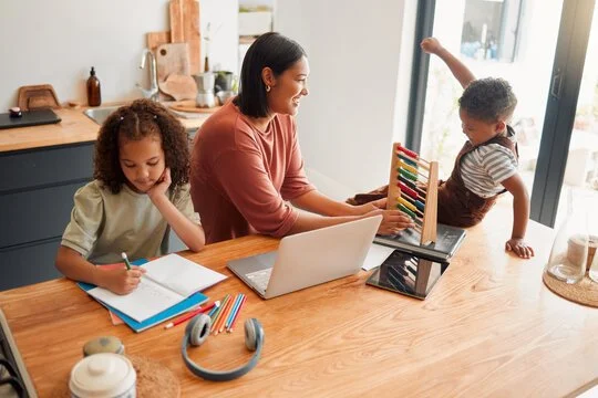 A woman and two children are sitting at a kitchen counter with a laptop, notebooks, and art supplies, while the boy performs a handstand near the window.