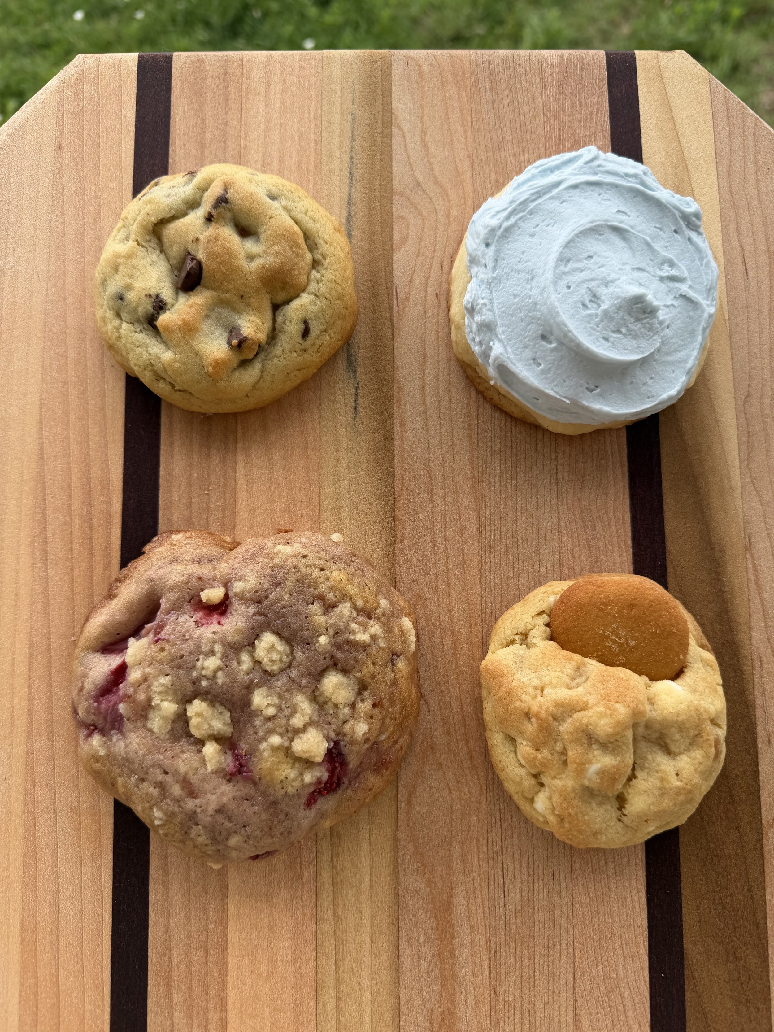 Image of four cookies on a wooden cutting board with green grass in the background. The cookies include a chocolate chip cookie, a frosted sugar cookie, a strawberry crumble cookie, and a banana pudding cookie with a Nila wafer on top.