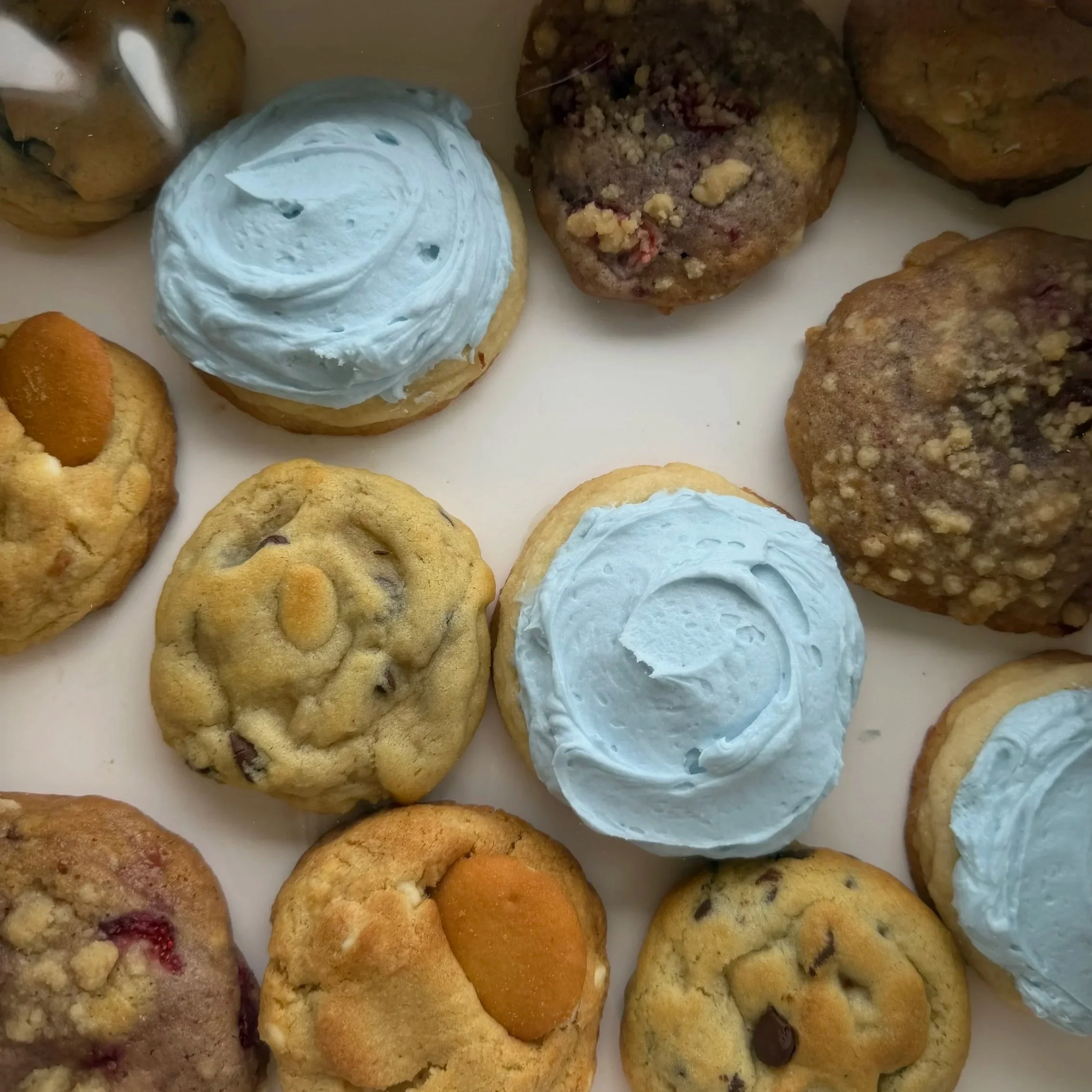 Assorted cookies including chocolate chip, berry, and sugar cookies, with some topped with frosting or nuts, on a white surface.