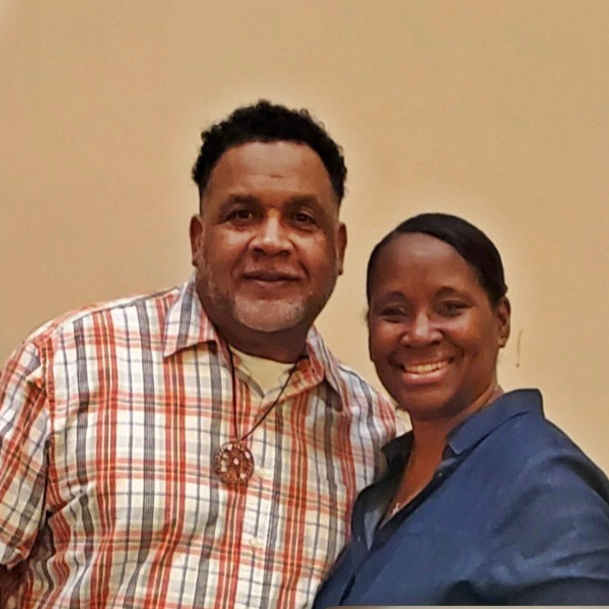 A man and woman smiling for a photo against a plain background. The man wears a plaid shirt and a necklace, and the woman wears a dark blue top.