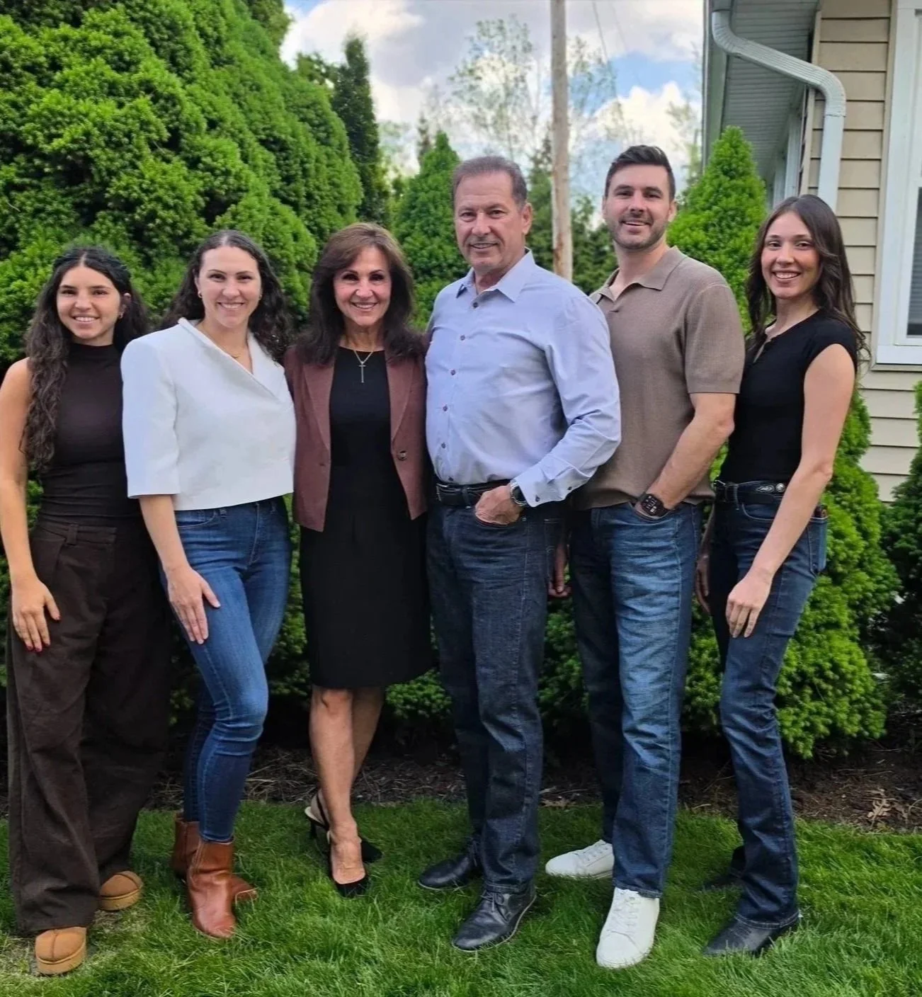 Group of six people standing outdoors in front of green bushes and house siding, smiling at the camera.