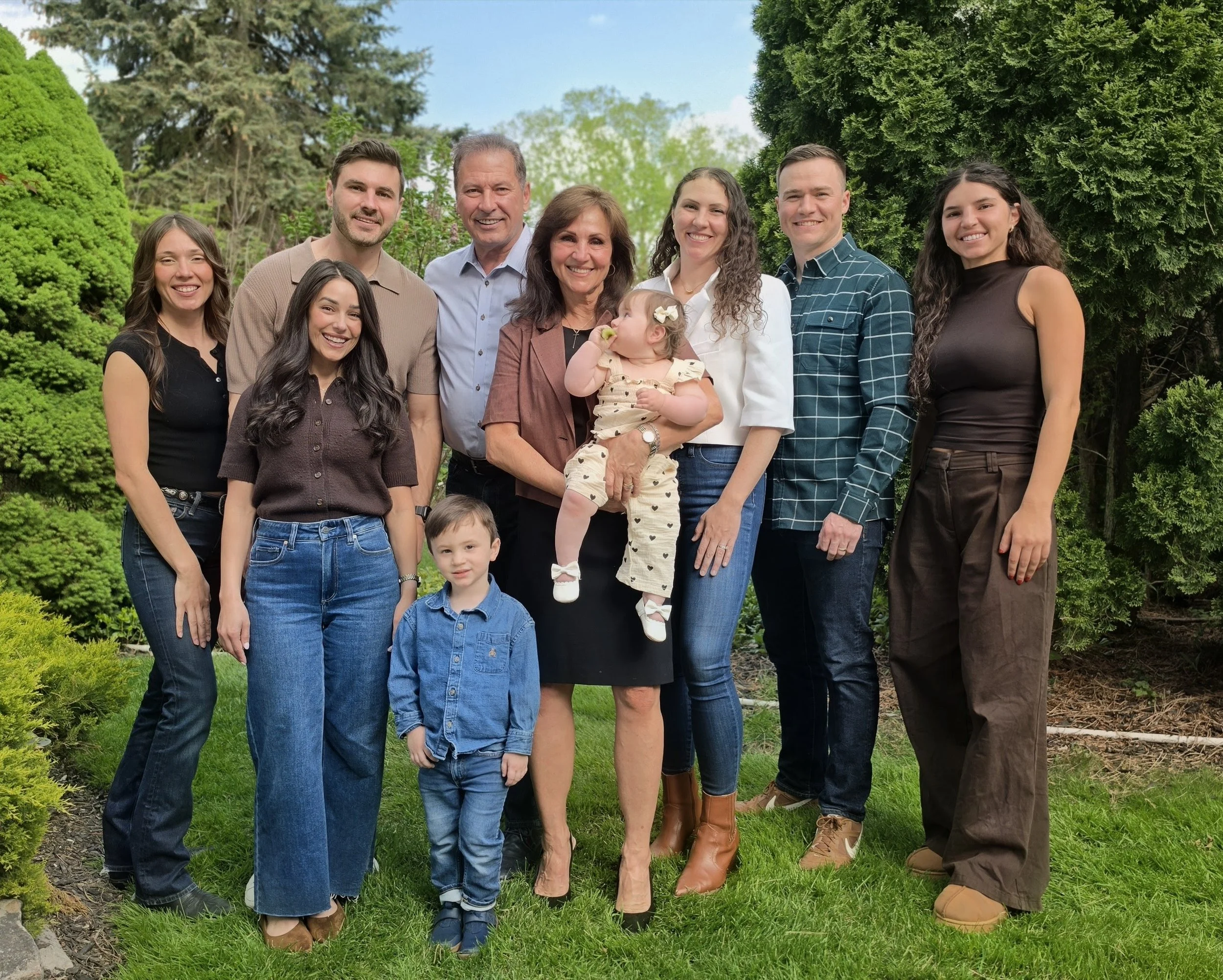 Group of nine people, including children, smiling and posing outdoors in a garden with lush green trees and bushes.