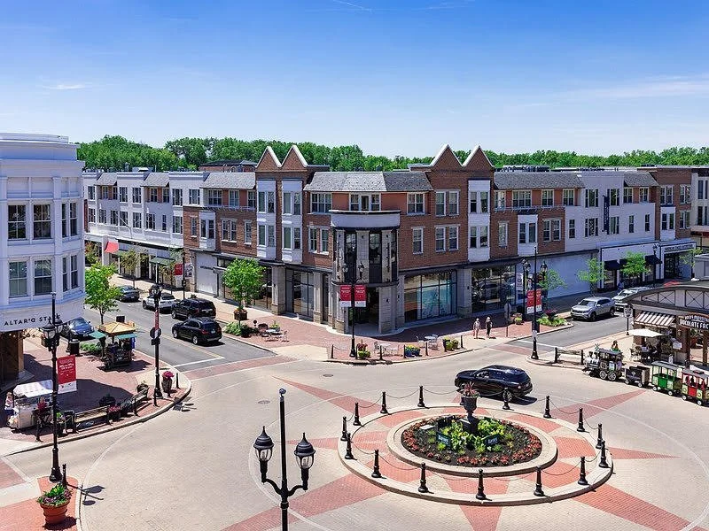 A small town square with a roundabout and flower bed in the center, surrounded by multi-story retail and apartment buildings, outdoor seating, and parked cars.