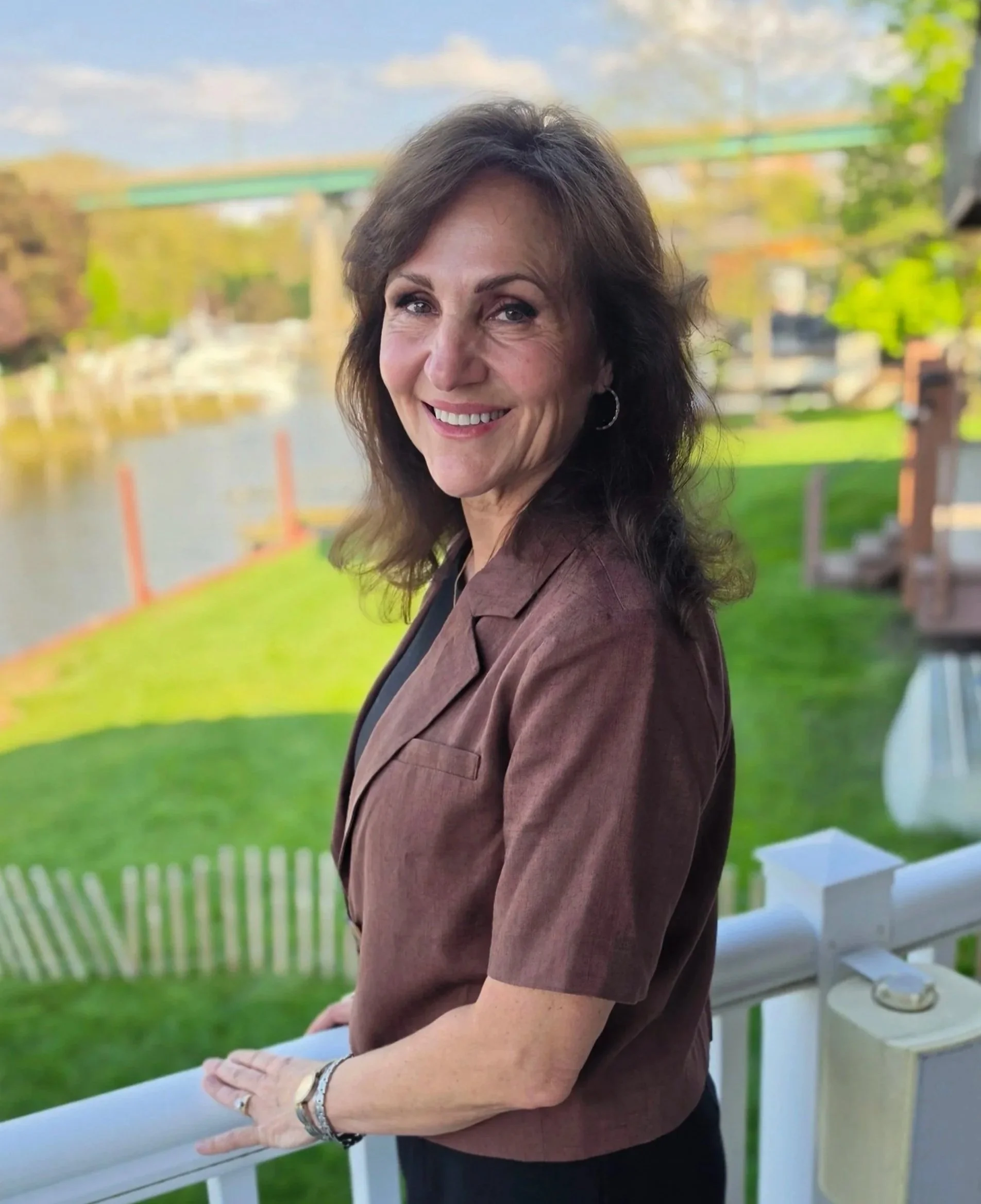 A woman smiling outdoors near a river, with a green bridge in the background, standing next to a white fence.