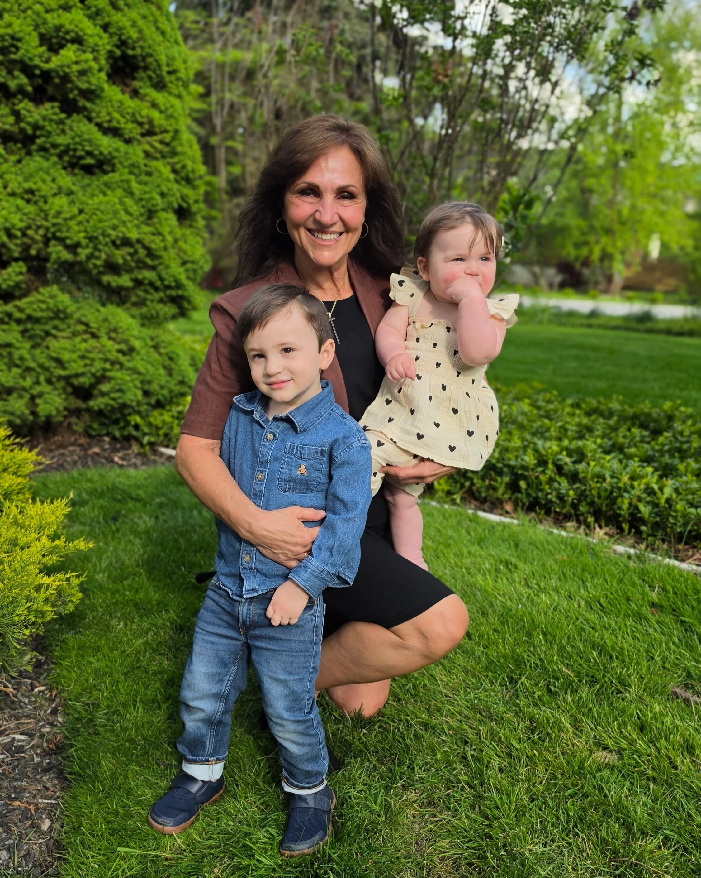 A woman holding a young girl and a young boy outdoors in a garden with greenery. The woman is smiling and wearing a brown blazer, the girl is wearing a beige dress with black hearts and has her thumb in her mouth, and the boy is wearing a blue denim jacket and has short hair.