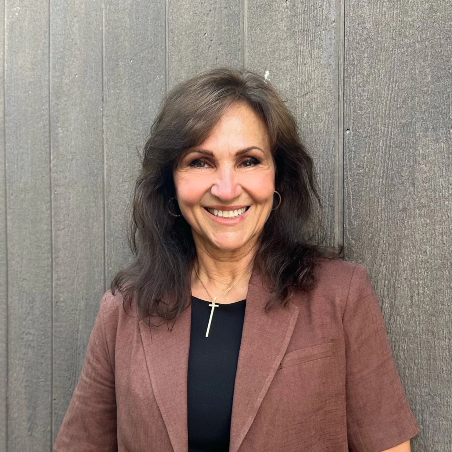 A woman with long, dark, wavy hair smiling and wearing a brown blazer, black top, hoop earrings, and a necklace with a cross pendant, standing against a gray wooden wall.