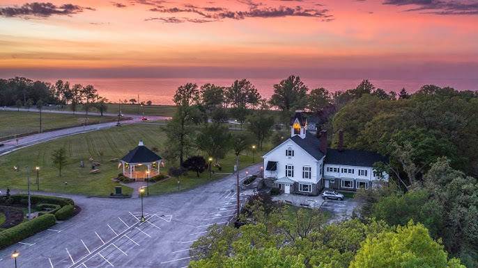 A scenic view of a small town or village with a church and a white house surrounded by trees, under a colorful sunset sky with pink, purple, and orange hues.
