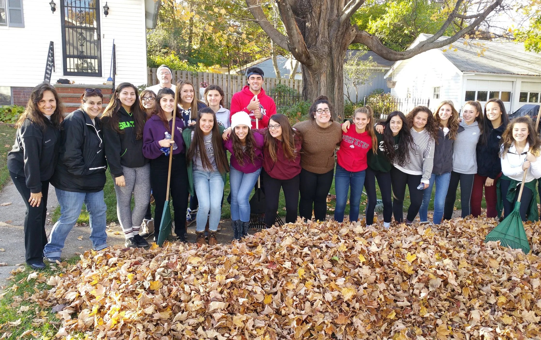 A group of people standing outdoors behind a large pile of fallen autumn leaves, smiling at the camera. They are in front of a house with a large tree overhead and other houses in the background. It is daytime with clear weather.