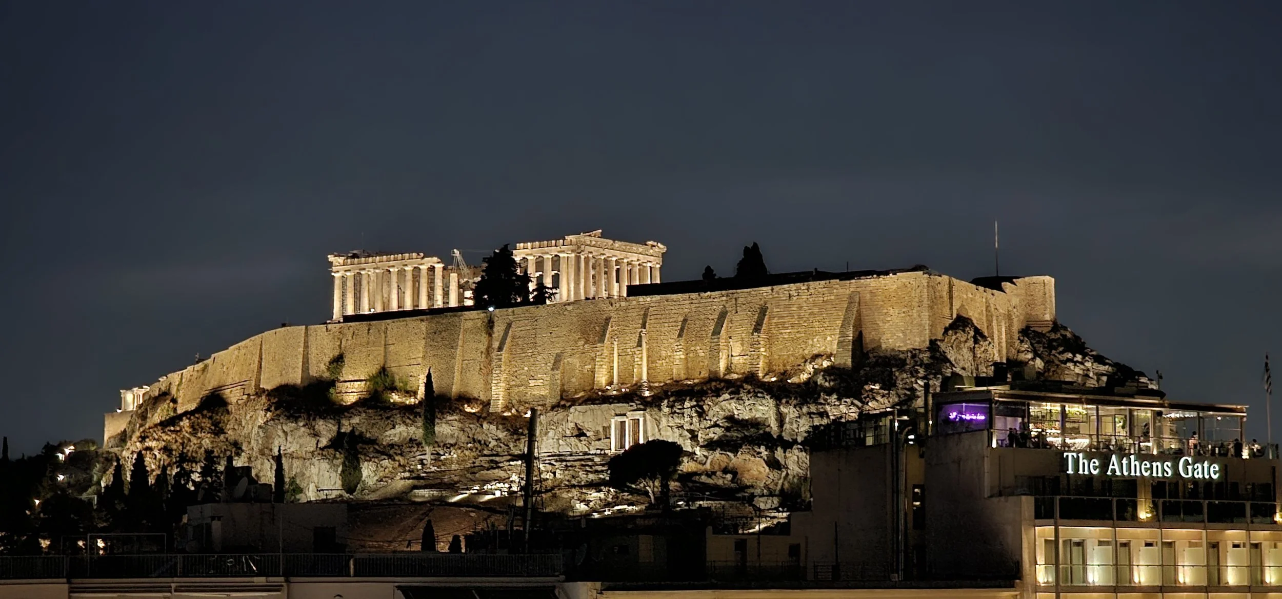Night view of the illuminated Parthenon atop the Acropolis in Athens, Greece, with the sign reading "The Athens Gate" visible in the foreground.