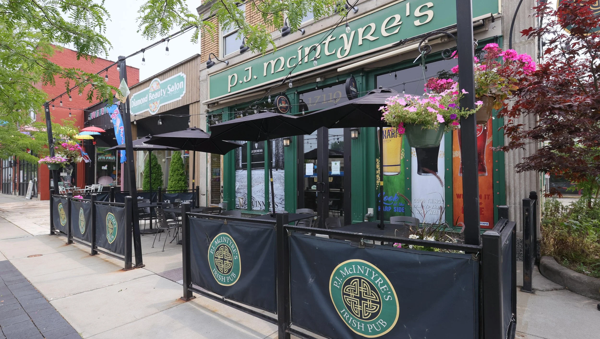 Exterior of P.J. McIntyre's Irish Pub with outdoor seating, black umbrellas, flower planters, and string lights, located on a downtown street lined with trees and building storefronts.