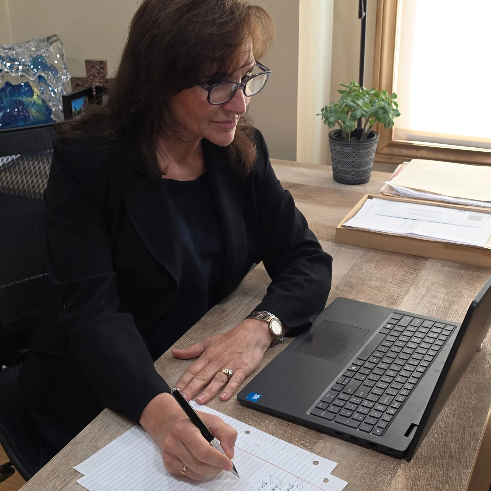 A woman with brown hair, glasses, wearing a watch and rings, sitting at a desk with a laptop, writing on a lined sheet of paper with a black pen. There is a potted plant with green leaves, some papers, and other items on the desk, and a window with natural light to her right.