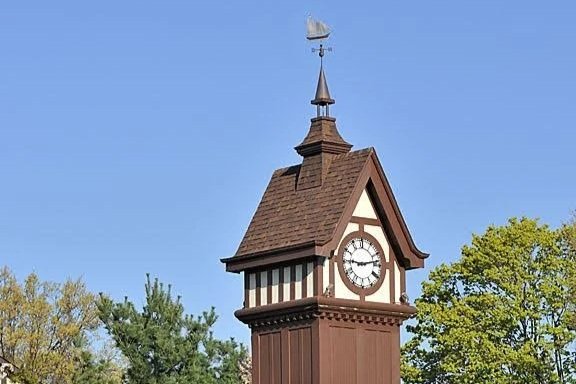 A clock tower with a weather vane on top, surrounded by trees against a clear blue sky.